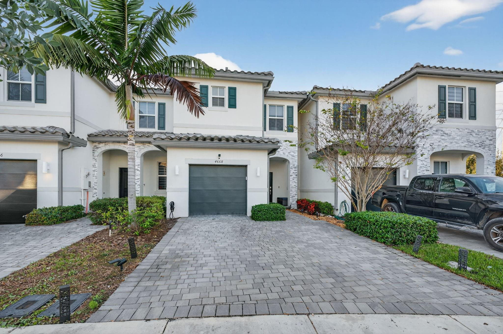 Two-story white townhouse with stone accents, paved driveway, and palm tree.