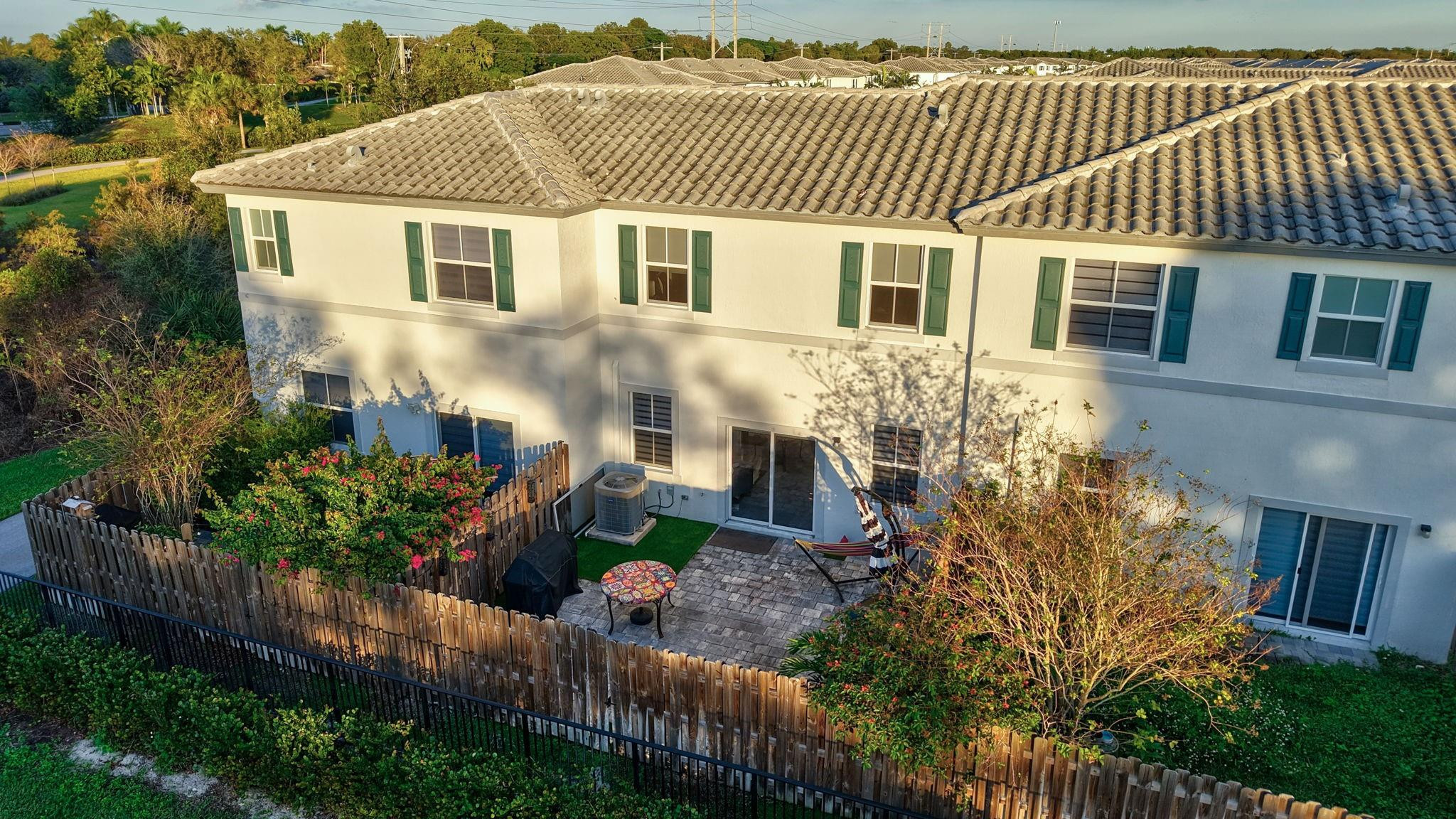 Rear of a two-story home with a fenced patio, pavers, and flowering shrubs.