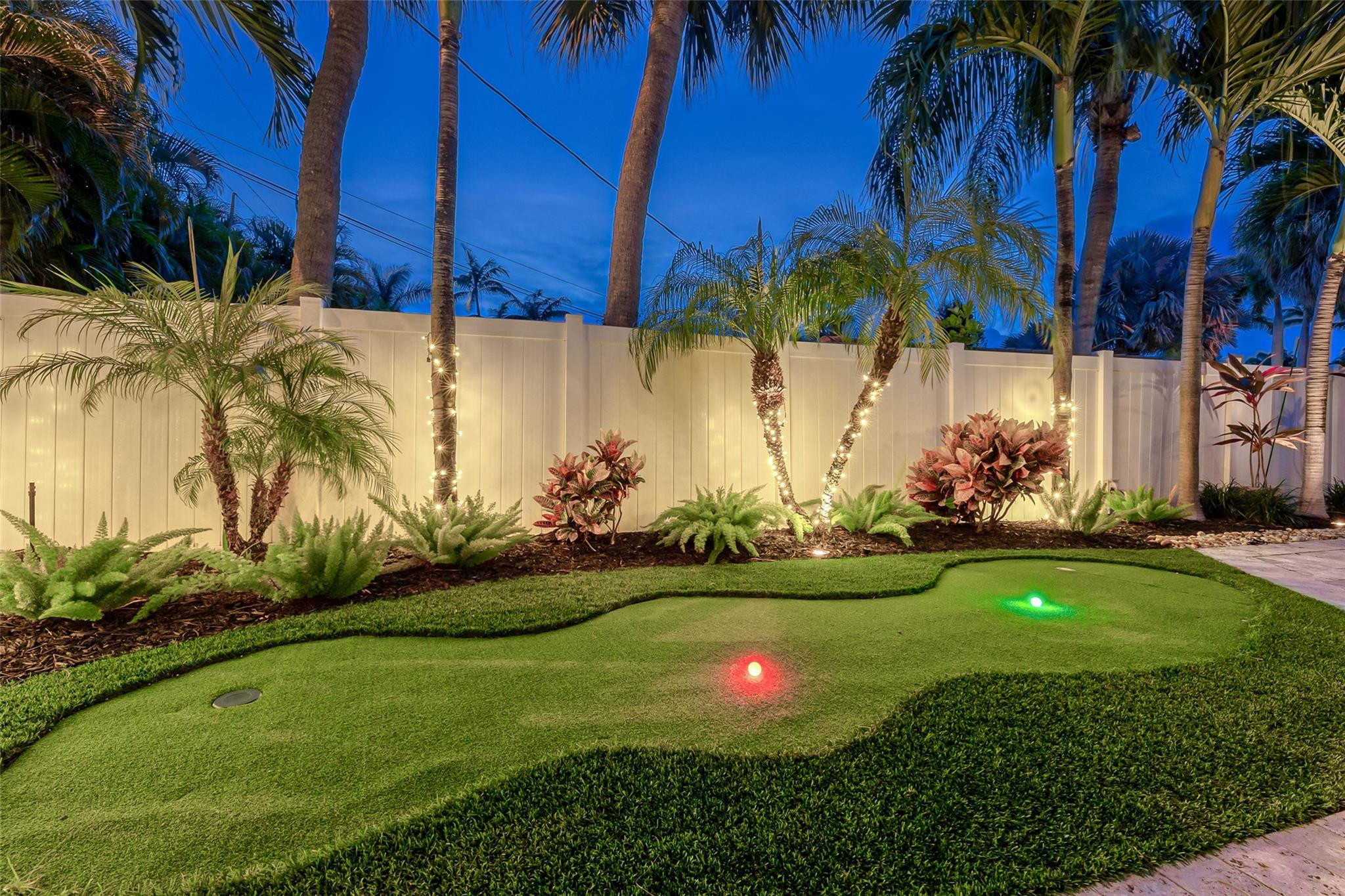 Backyard putting green with illuminated holes, tropical palms, and a white fence at dusk.