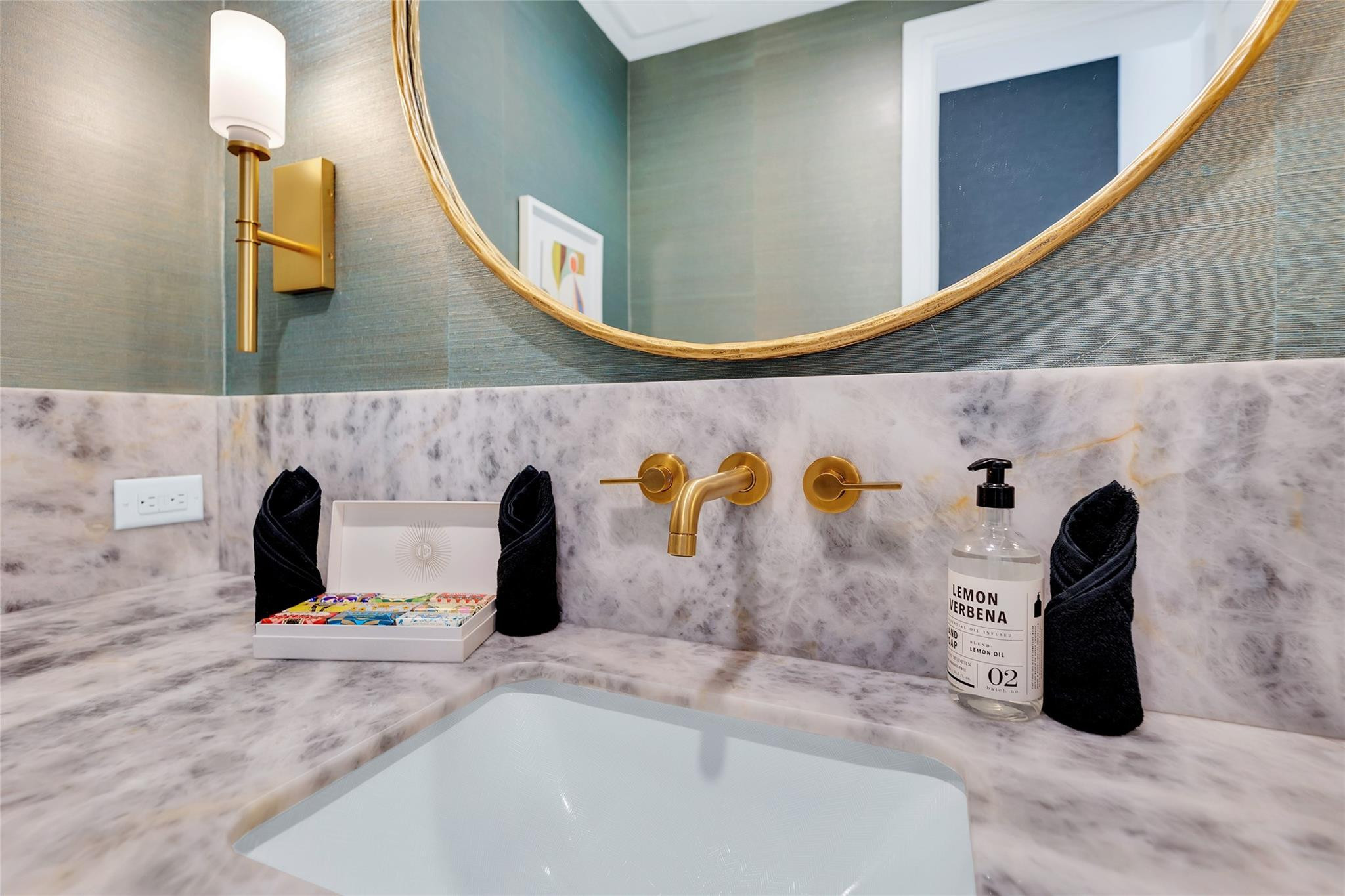 Bathroom vanity with marble surfaces, gold fixtures, round mirror, black towels, and a lemon verbena soap bottle.