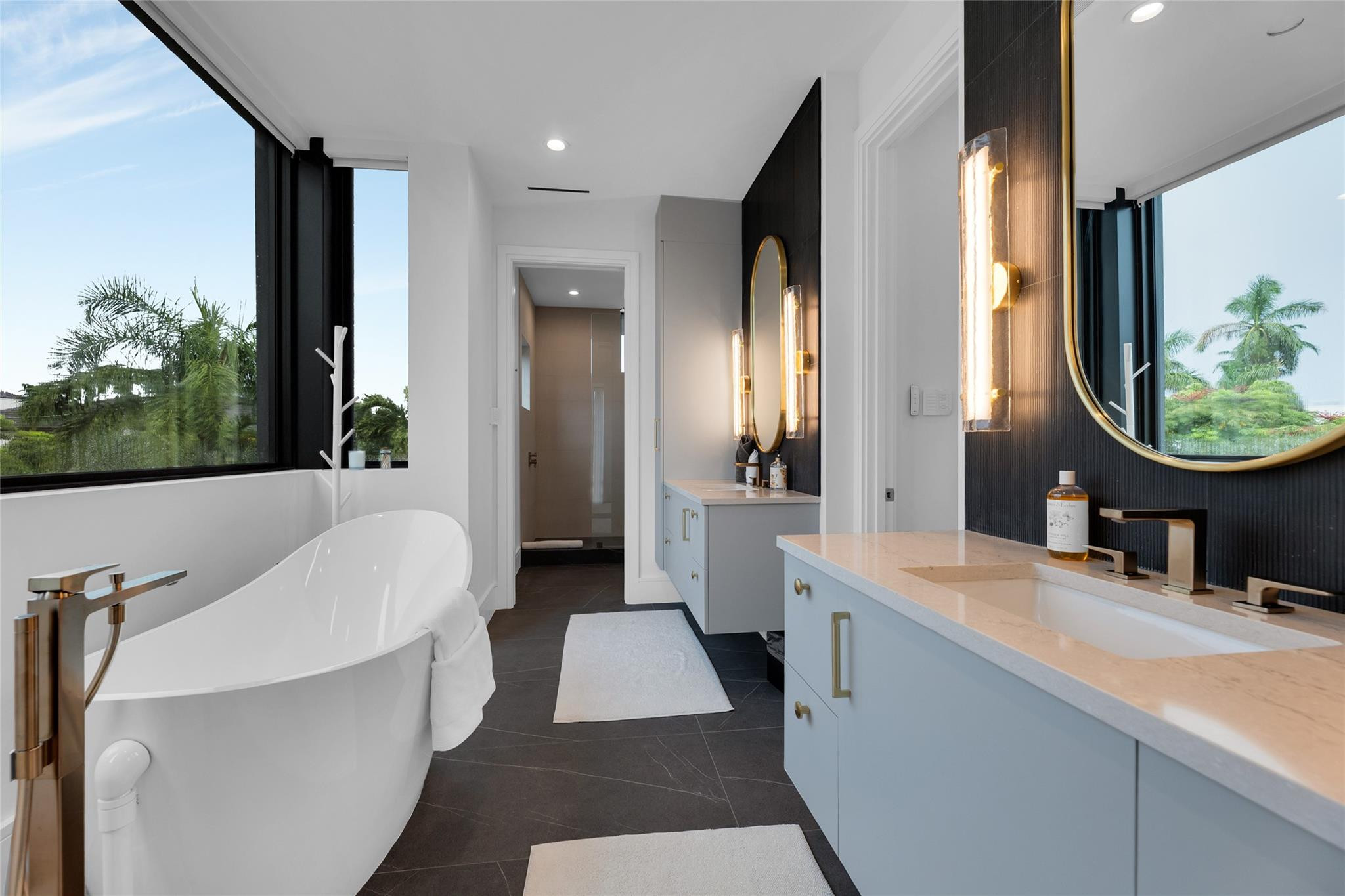 Modern bathroom with freestanding tub, palm-view windows, and pale-blue double vanity.