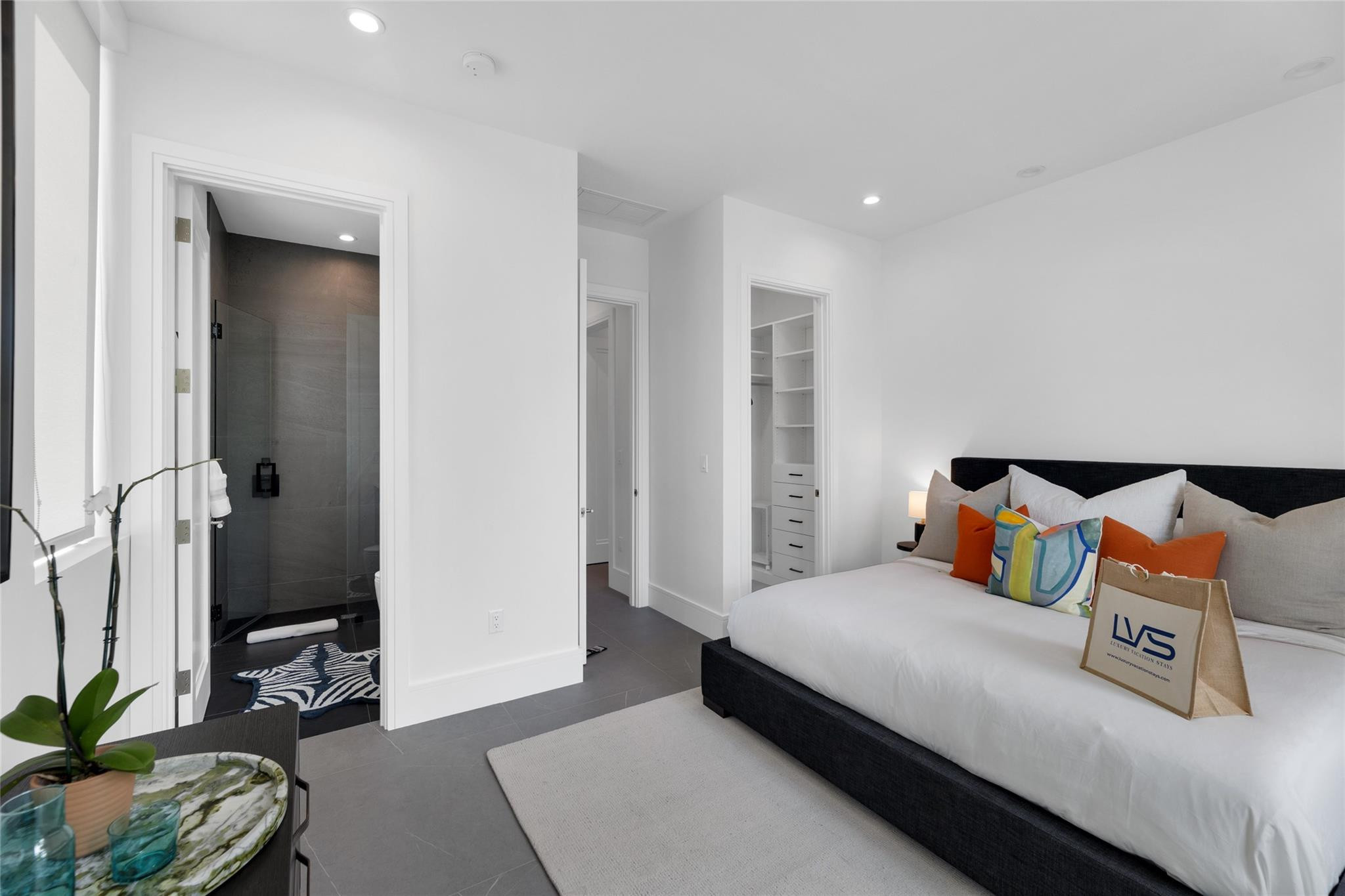 Staged bedroom with white bedding, colorful accent pillows, and an adjoining dark-tiled shower.