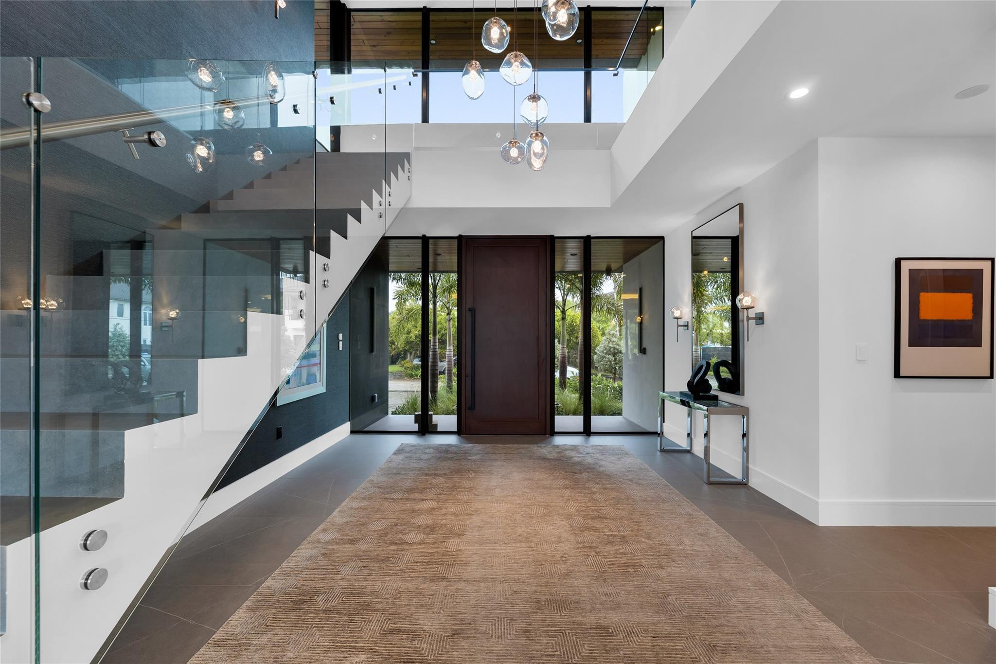 Modern entry foyer with a tall dark wood door, glass staircase railing, pendant lights, and a large rug.