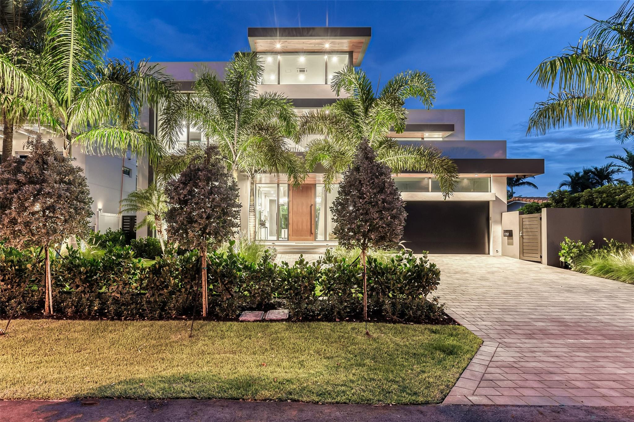 Modern three-story home with a glass entry, wide driveway, and palm-lined landscaping at dusk.