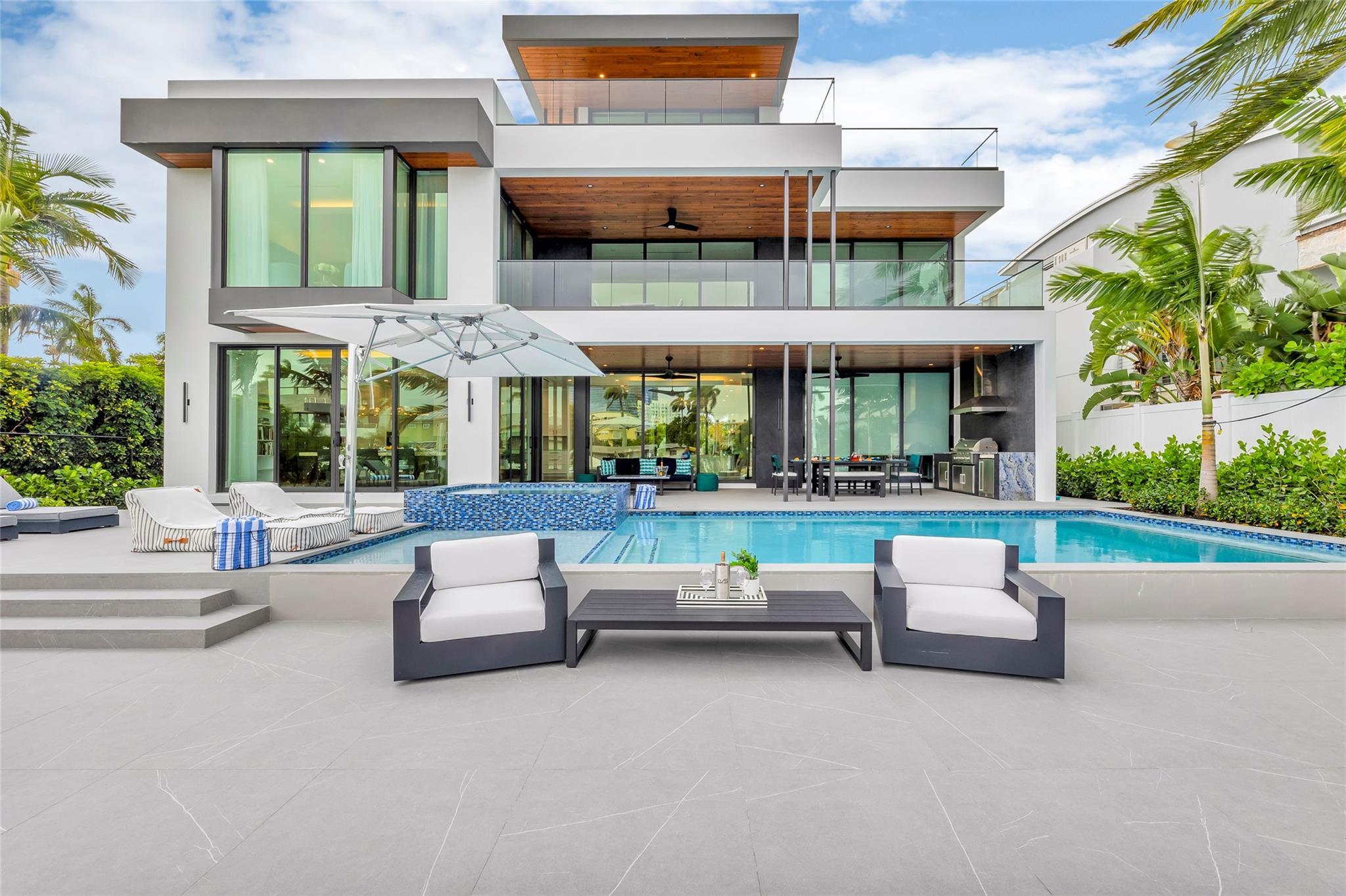 Modern glass-fronted home overlooking a swimming pool and furnished patio.