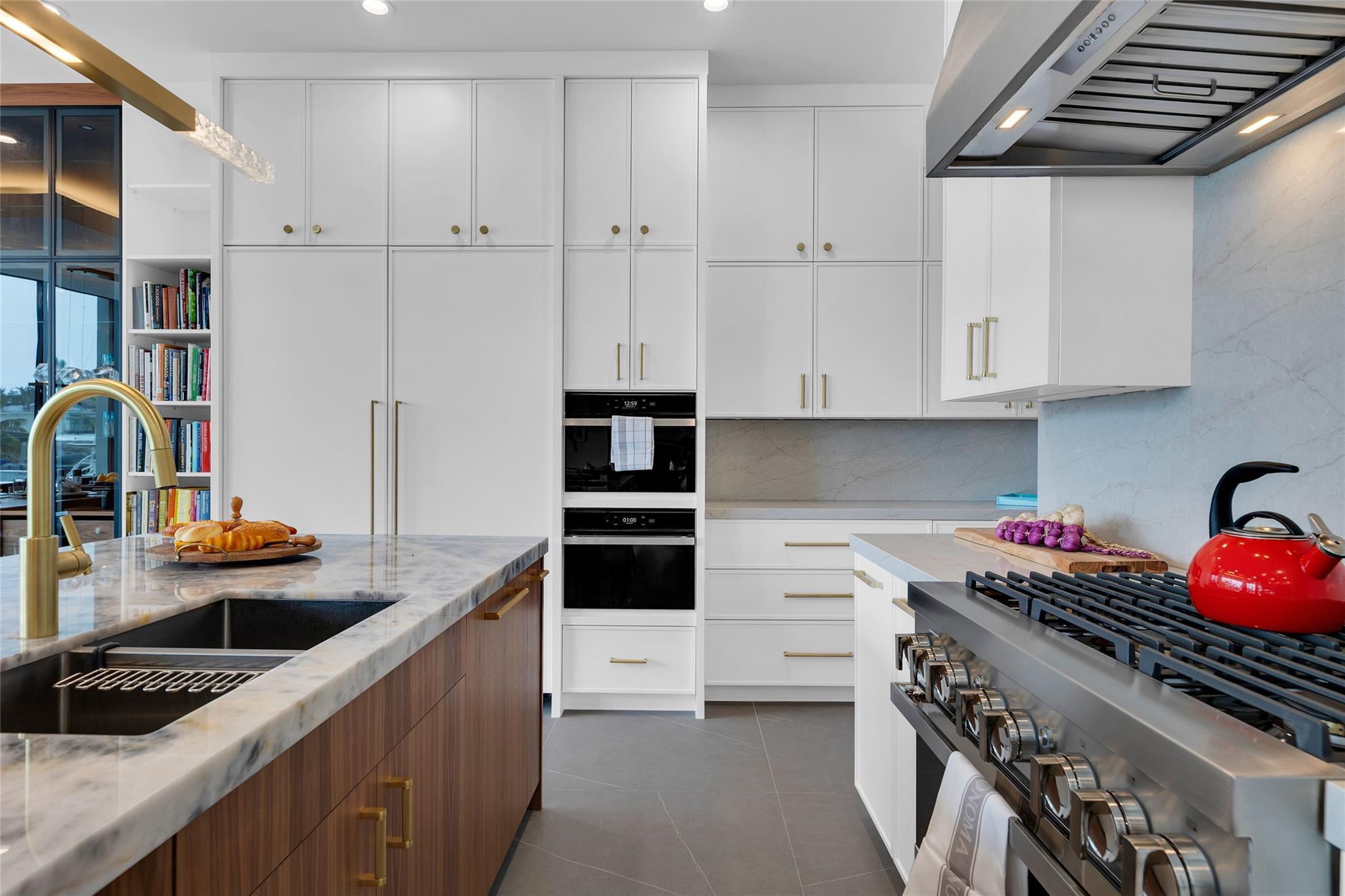 Modern kitchen with a marble island, white cabinets, double ovens, and a stainless steel range with red kettle.
