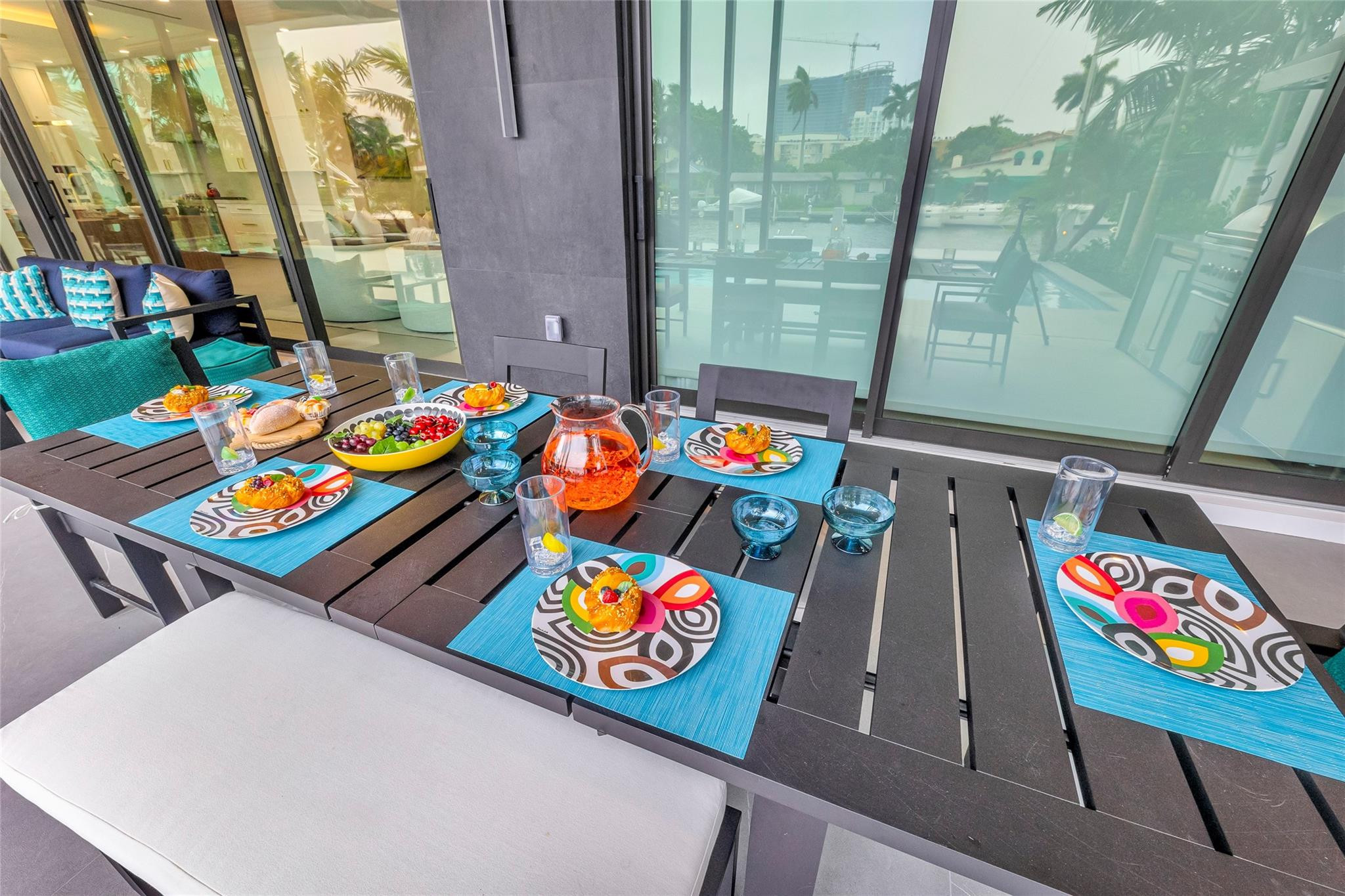 Patio dining table set with colorful plates and fruit, overlooking a canal through glass doors.