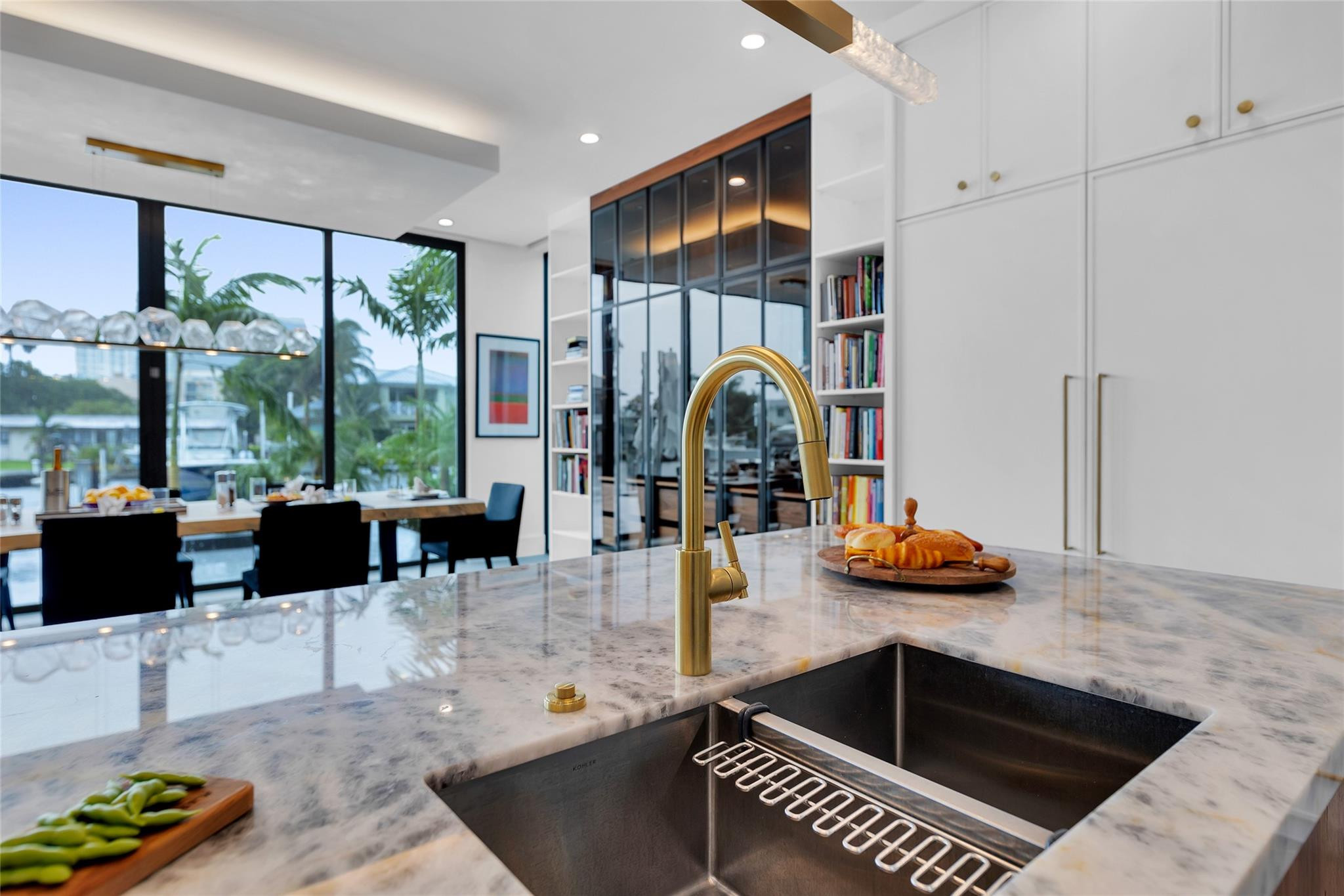 Modern kitchen with marble island, brass faucet, and large windows overlooking a palm-lined waterway.