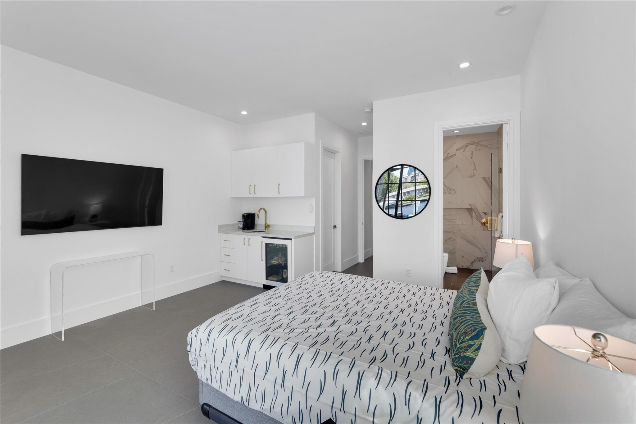 Modern bedroom with patterned bedding, a wall-mounted TV, compact kitchenette, and open marble-look bathroom.