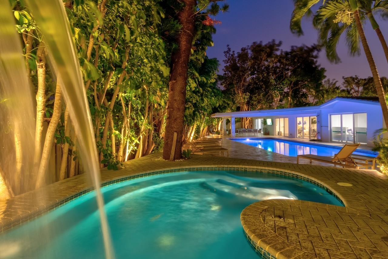 Modern home beside an illuminated pool and curved spa, framed by tropical foliage and palm trees.