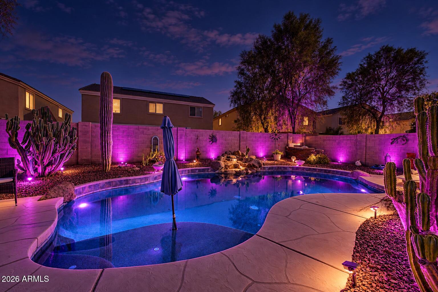 Backyard pool at night with vivid purple lighting, desert cacti, and curved stone decking.