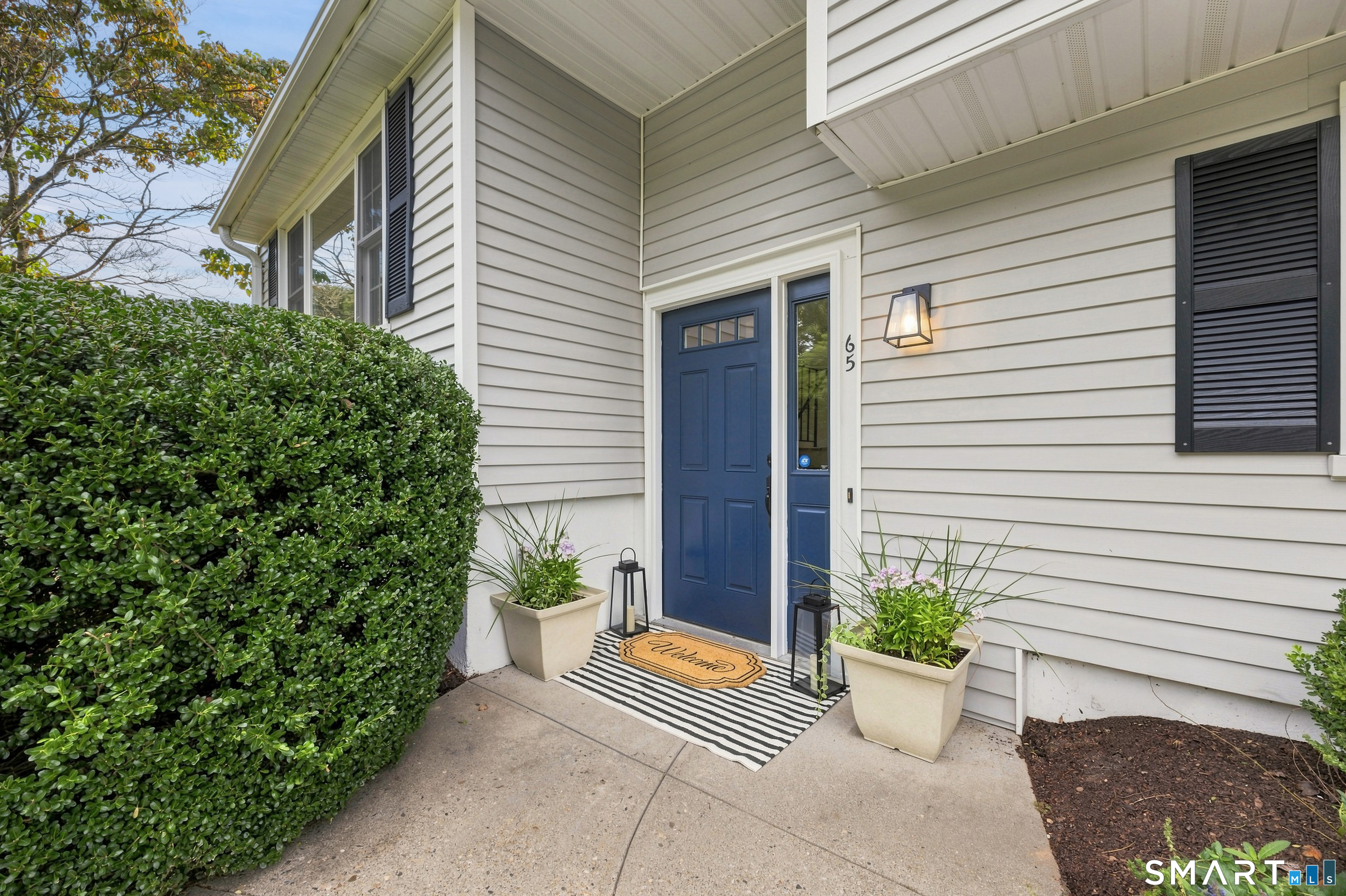 House entrance with a navy blue door, potted flowers, striped rug, and trimmed hedge.