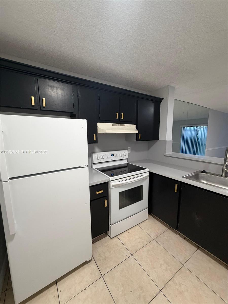 Compact kitchen with dark cabinets, white appliances, light counters, and beige tile flooring.