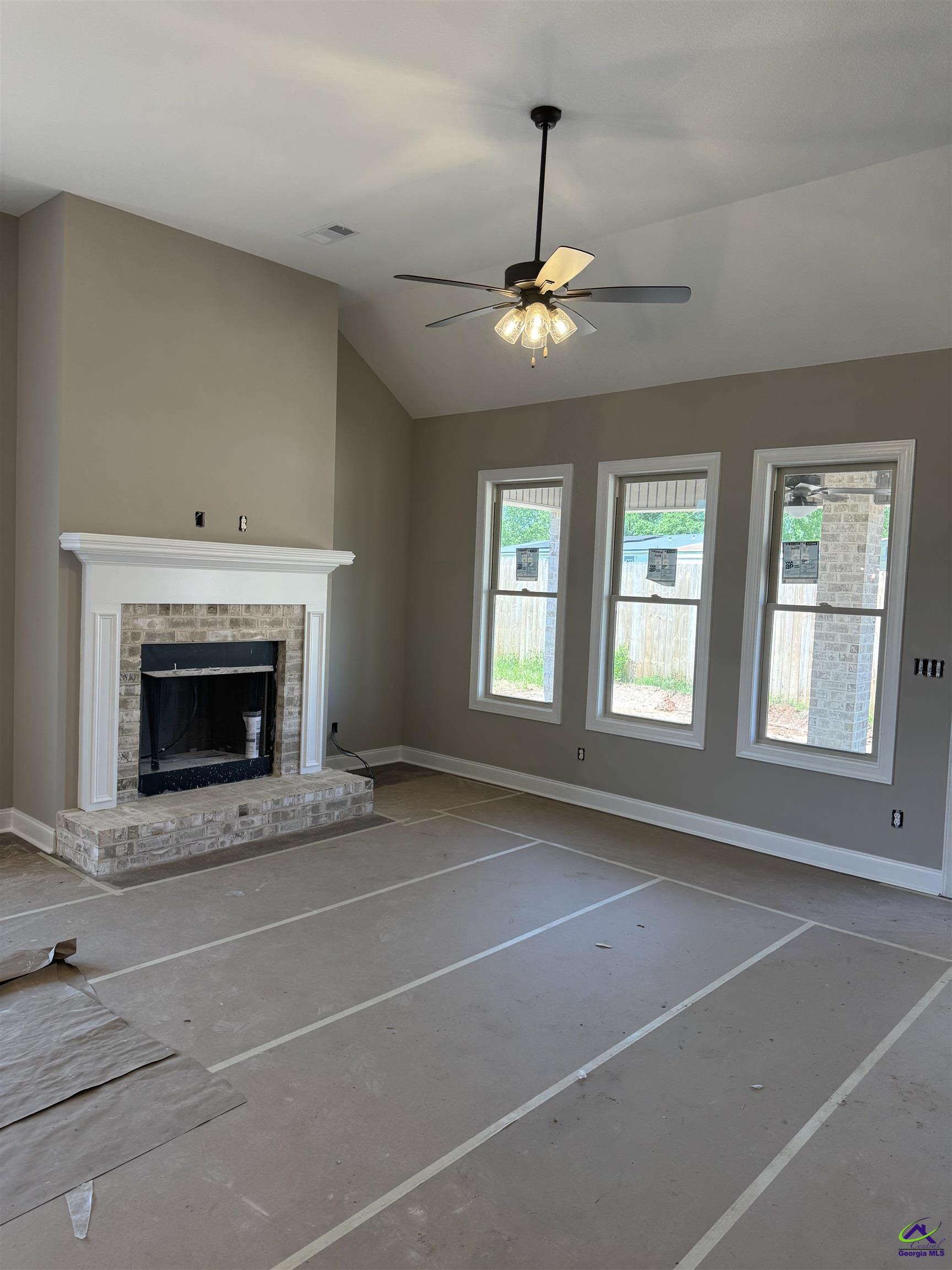 Unfinished living room with a brick fireplace, ceiling fan, and three tall windows.