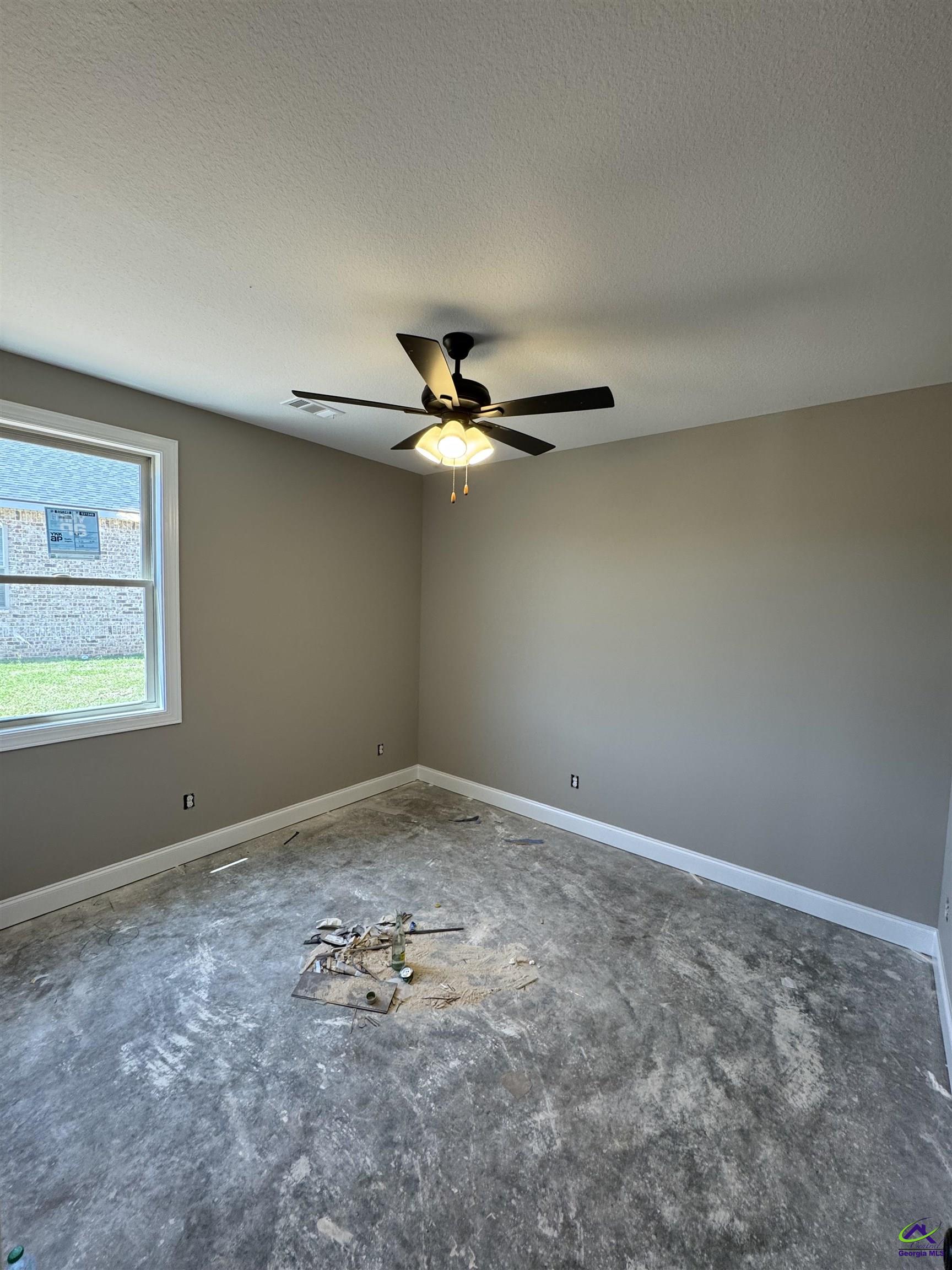 Empty room with a ceiling fan, window, and dusty floor scattered with renovation debris.