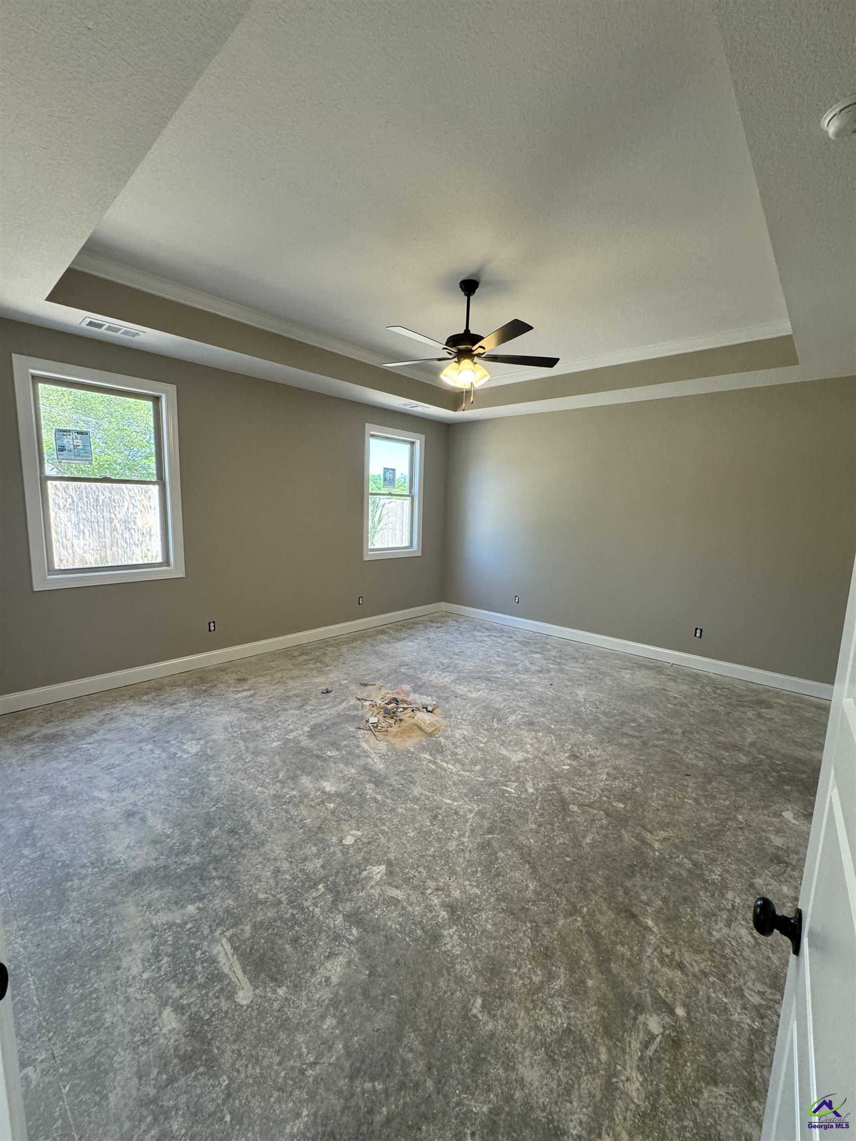 Unfinished vacant room with a tray ceiling, ceiling fan, two windows, and debris on the floor.