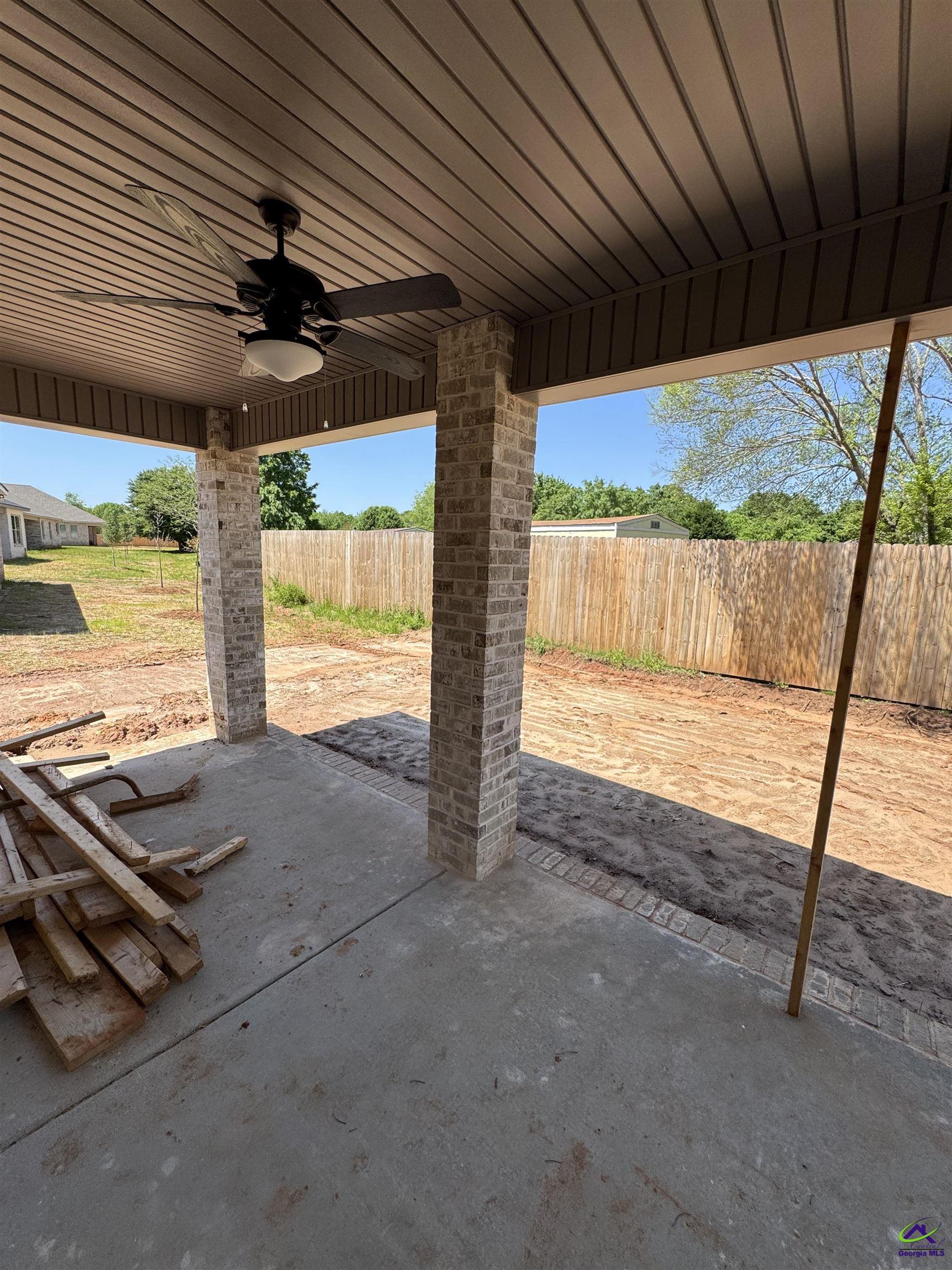 Covered concrete patio with brick columns and ceiling fan overlooking an unfinished fenced yard.