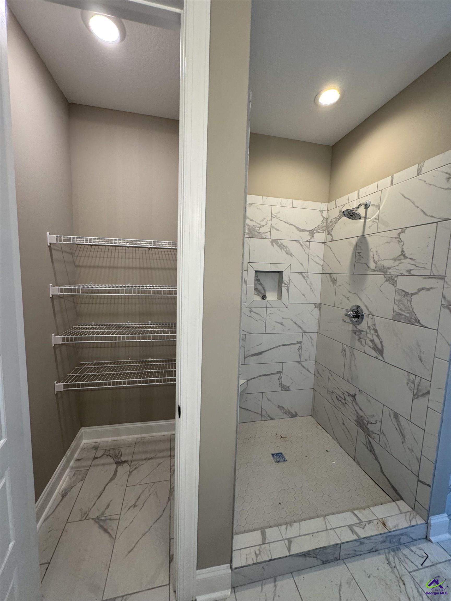 Bathroom walk-in shower with marble-look tile beside an open closet with wire shelving.