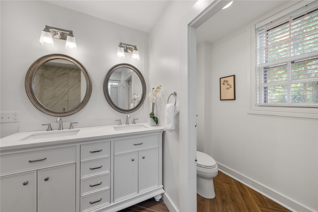 Bathroom with a white double-sink vanity, round mirrors, and a window beside the toilet.