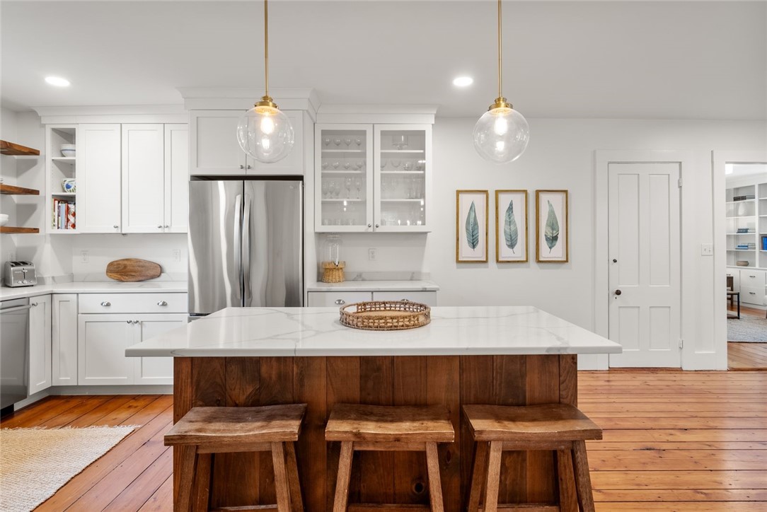 Bright white kitchen with a dark wood island, three stools, glass-front cabinets, and warm plank flooring.