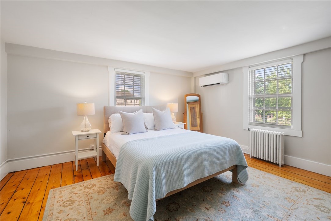 Bedroom with a white and pale blue bed, honey-toned hardwood floors, and two windows with blinds.