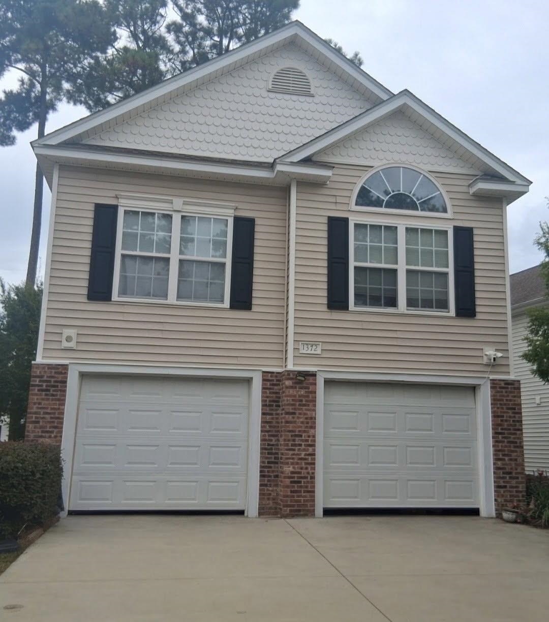 Beige two-story home with twin garage doors, brick lower facade, and a broad concrete driveway.