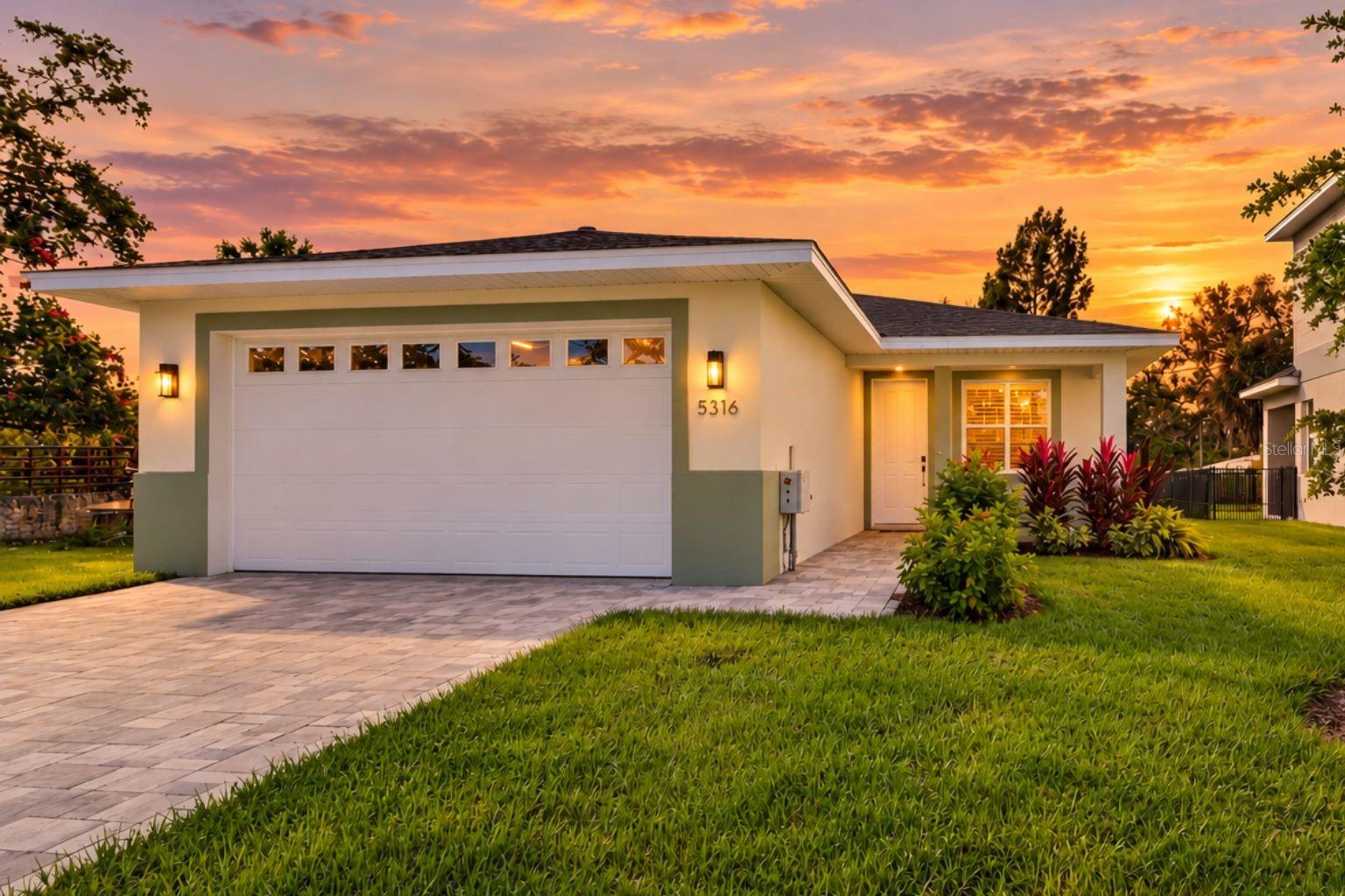 Single-story home with a wide garage and landscaped front yard beneath a vivid sunset sky.