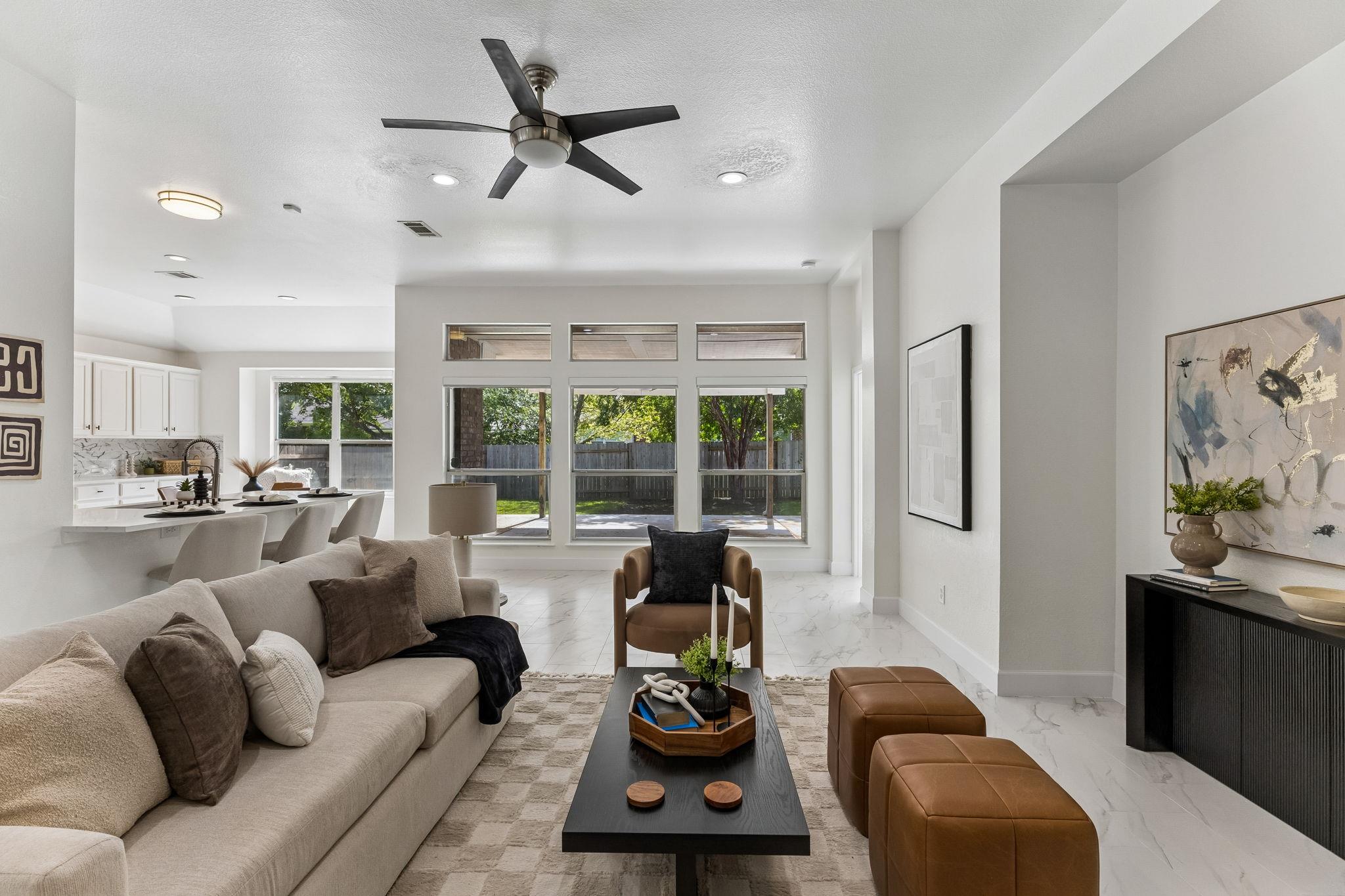Staged living room with a cream sectional, tall backyard windows, and an open kitchen.