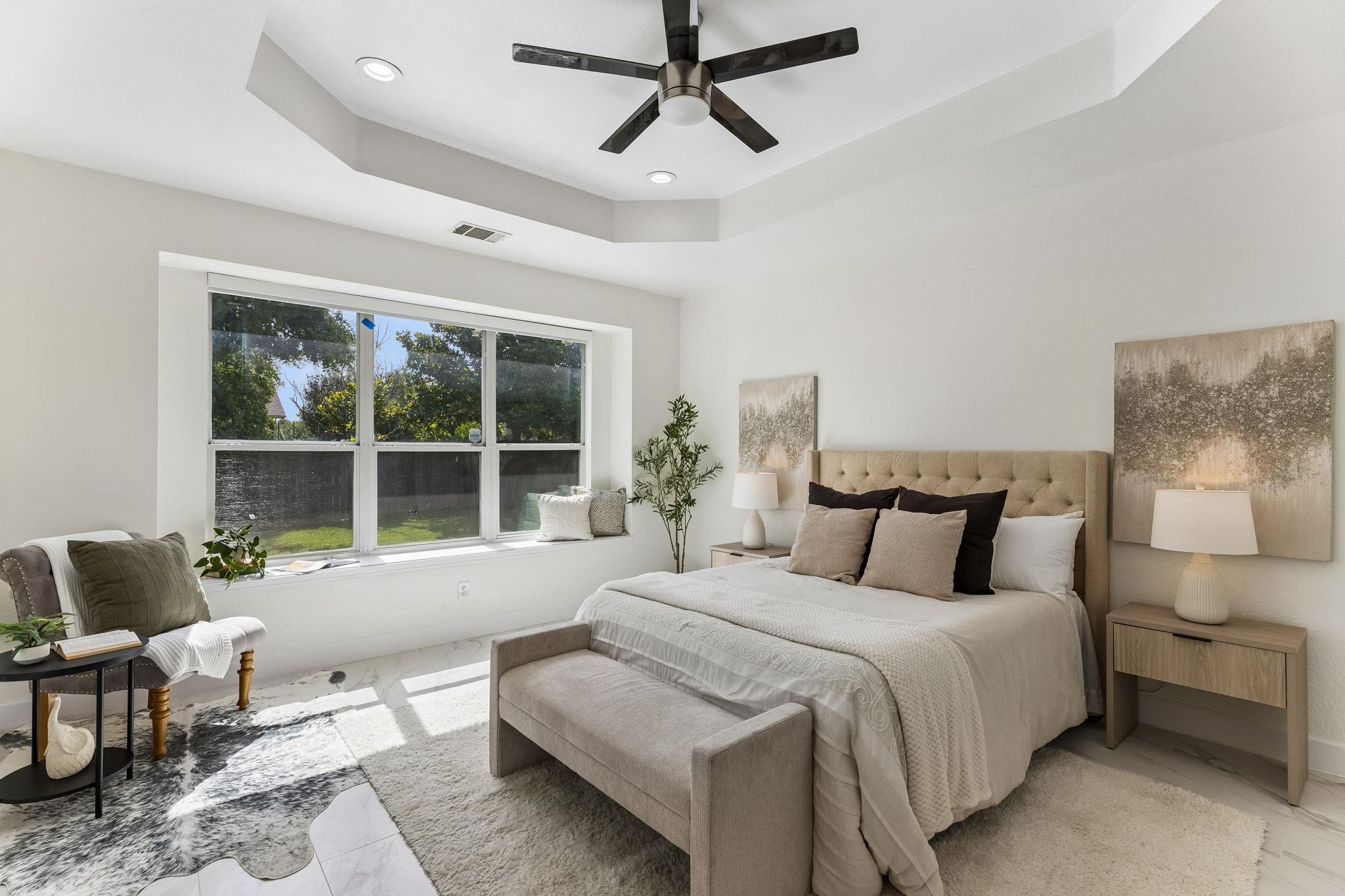 Staged bedroom with a tufted bed, tray ceiling and large garden-facing window.
