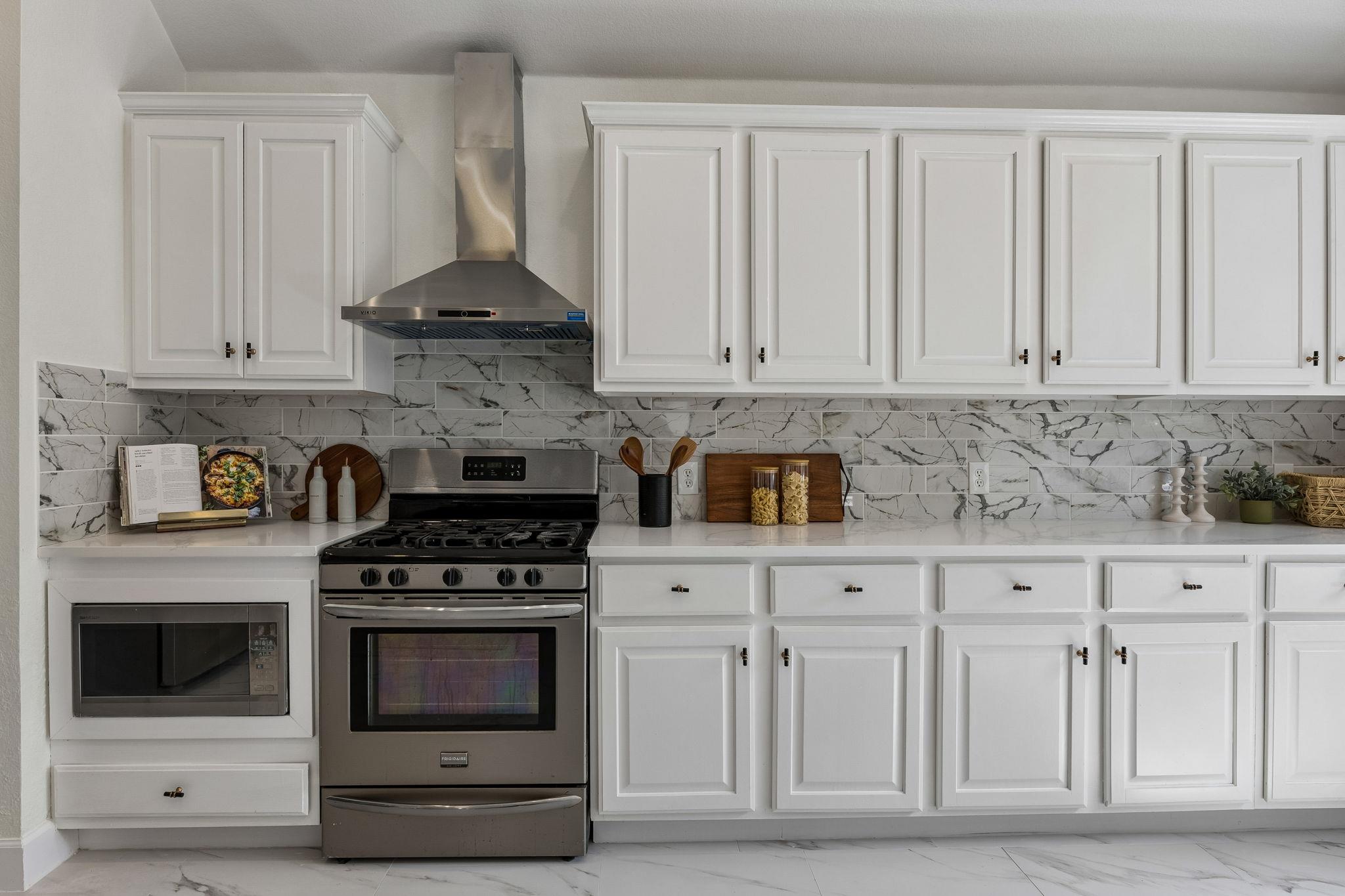 Kitchen with white cabinets, marble-patterned backsplash, and stainless steel range beneath a chimney hood.