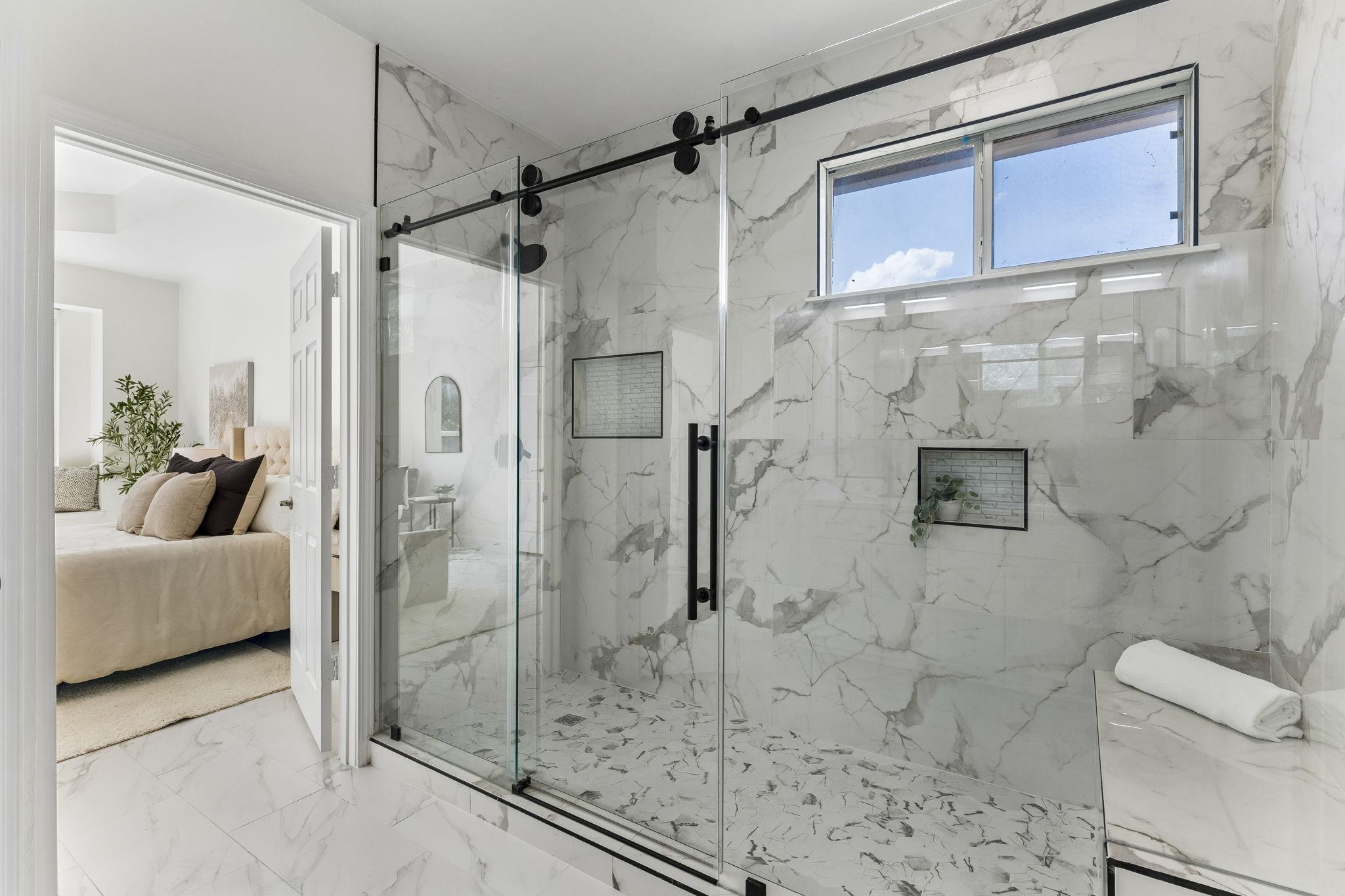 Marble-tiled walk-in shower with black hardware beside a furnished bedroom.
