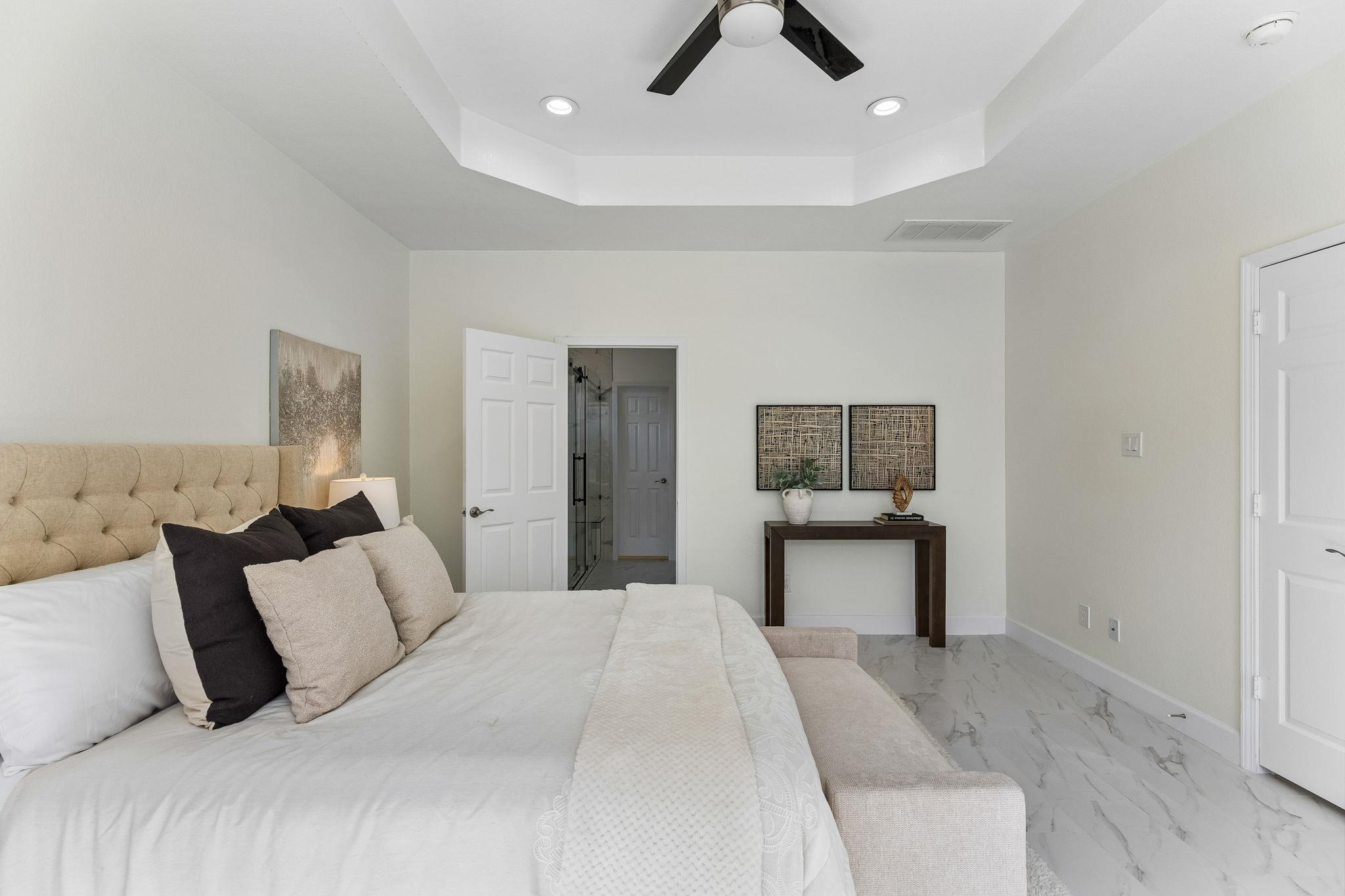 Staged bedroom with a tufted upholstered bed, tray ceiling, and marble-look flooring.