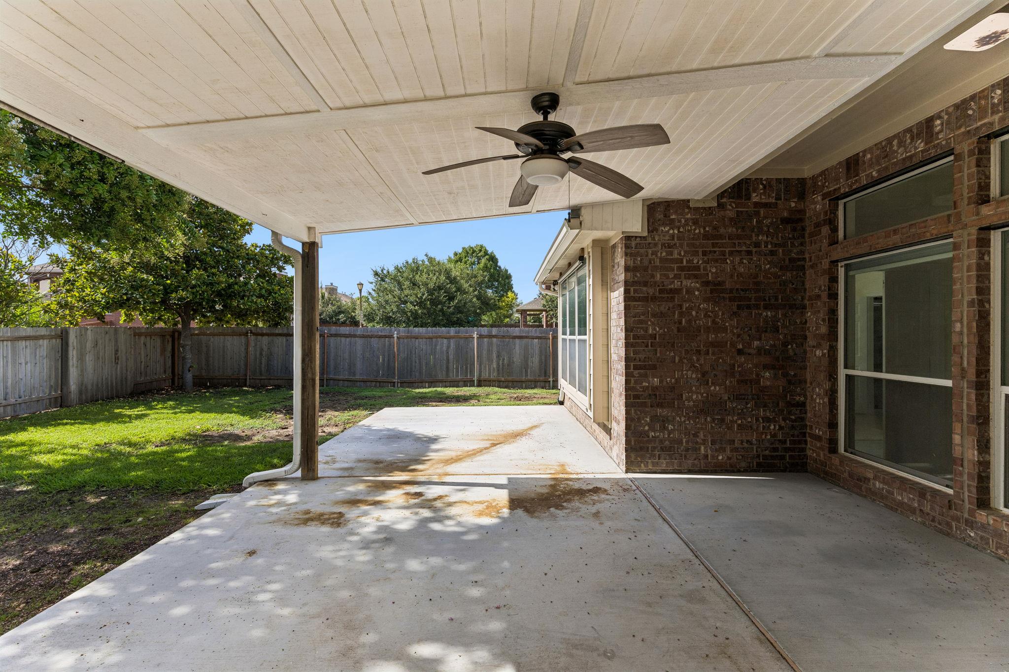 Covered backyard patio with a ceiling fan beside a fenced lawn and brick house.