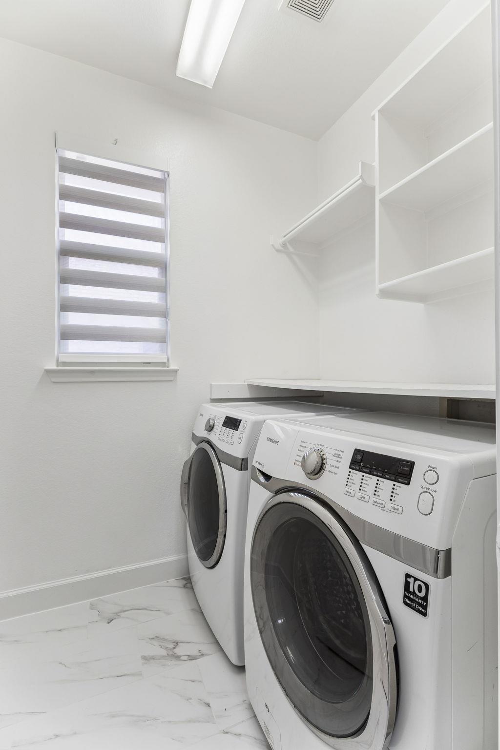 Laundry room with front-loading washer and dryer, white shelving, and a window with striped blinds.