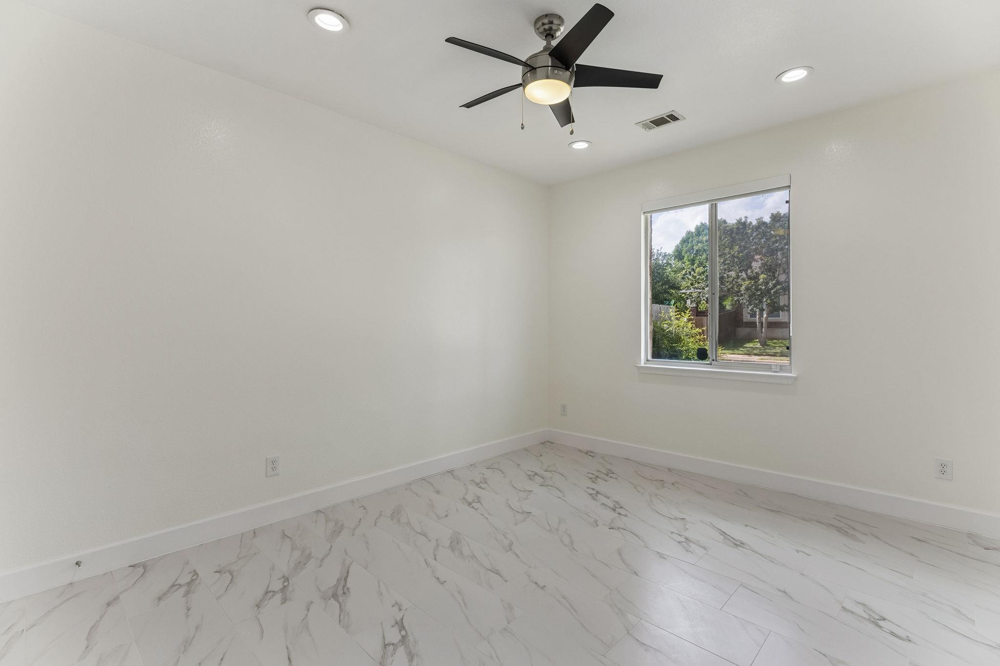 Empty room with marble-look tile flooring, a ceiling fan, and a window overlooking trees.