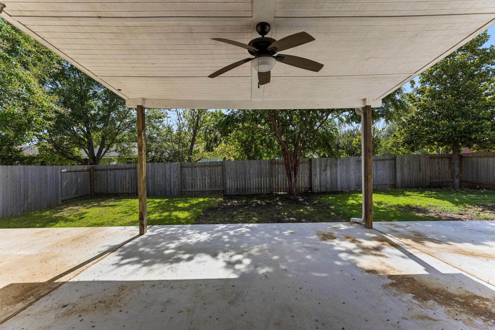 Covered backyard patio with ceiling fan overlooking a fenced, tree-shaded lawn.