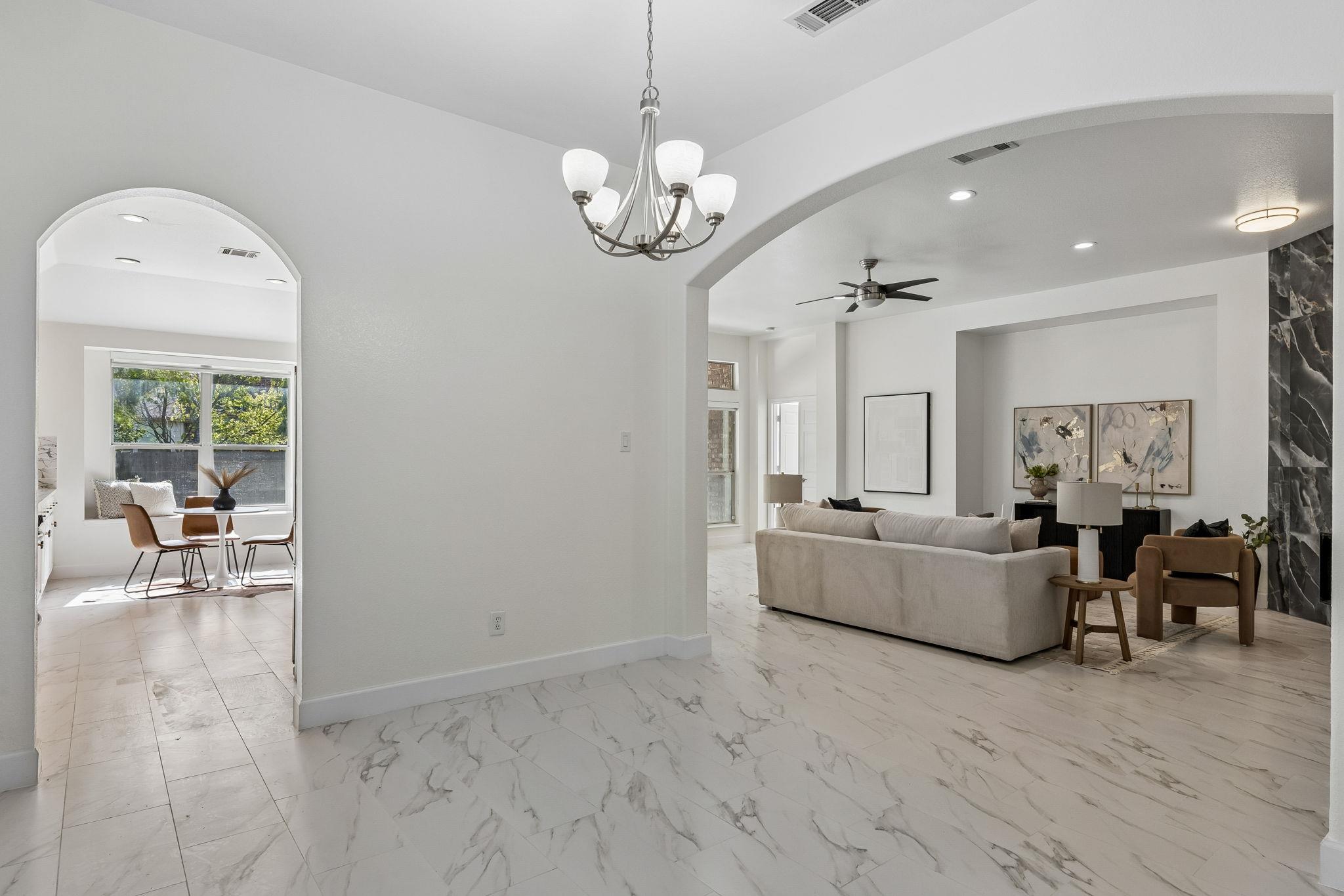 Staged living room with an arched opening to a sunny dining nook and marble-look tile floors.
