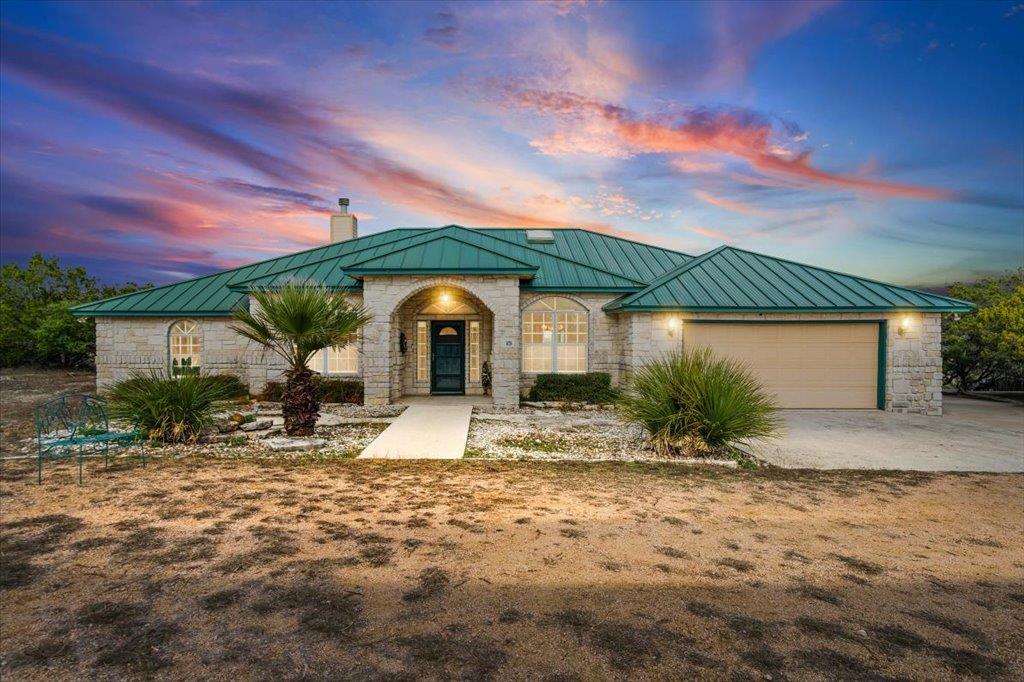 Stone house with a green metal roof and two-car garage beneath a colorful sunset sky.