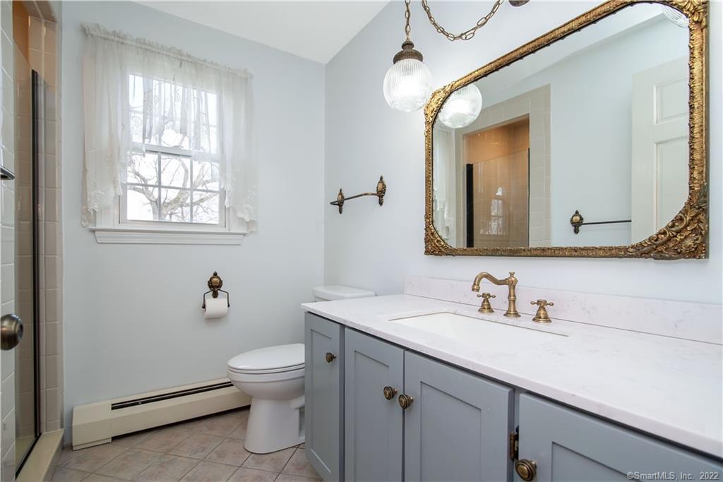 Bathroom with gray vanity, white countertop, ornate gold-framed mirror, and window with sheer curtains.