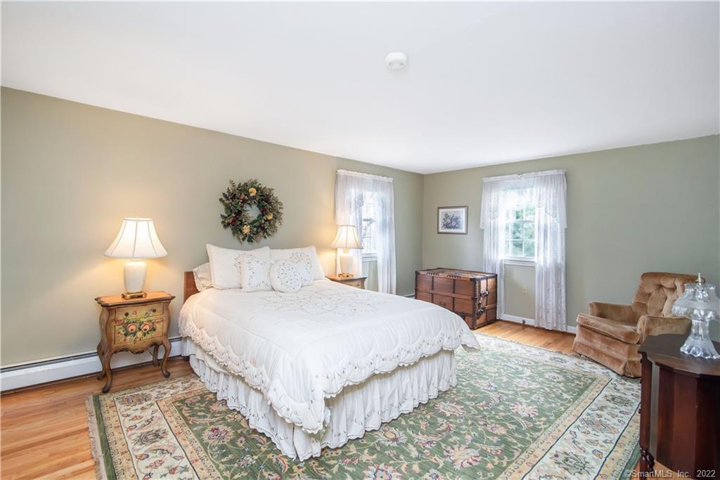 Bedroom with a white bed, floral rug, antique chest, and two bright curtained windows.
