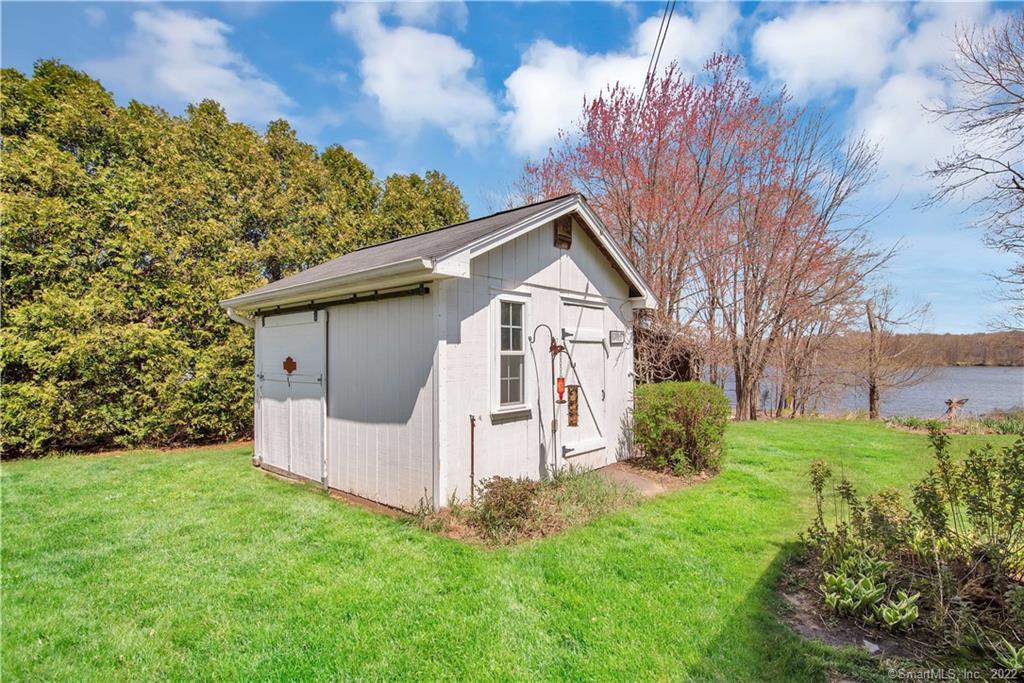 White detached shed on a green lawn, with trees and water behind it.
