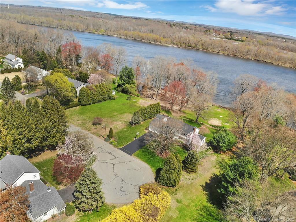 Aerial view of homes and lawns beside a broad river, with a curved road in the foreground.