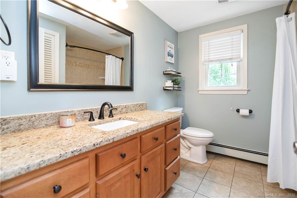 Bathroom with a granite vanity, warm wood cabinets, large mirror, tiled floor, and window with white blinds.