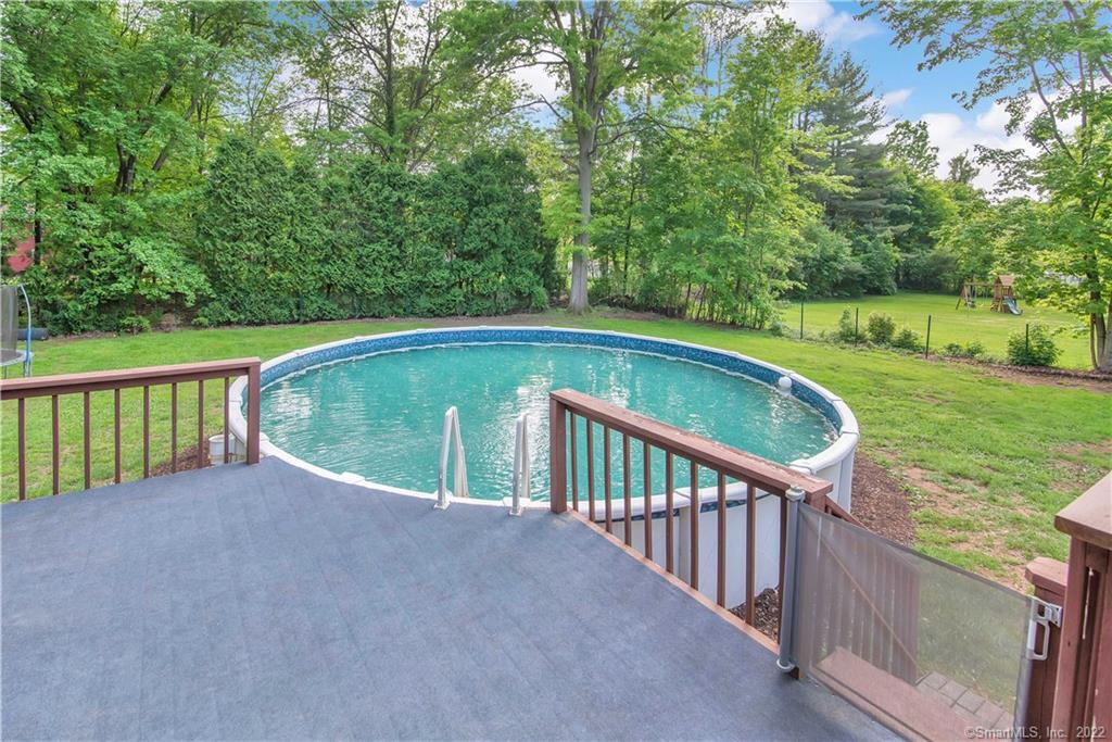 Backyard pool beside a gray deck, with a green lawn, mature trees, and a playset.