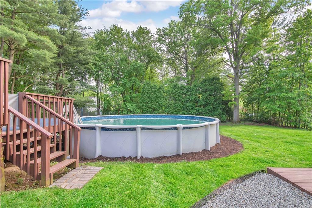 Above-ground backyard pool beside a wooden deck, surrounded by lawn and mature trees.