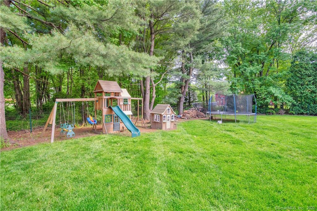 Grassy backyard with a wooden playset, small playhouse, and trampoline beneath mature trees.