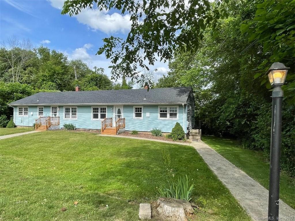 Light-blue single-story home with two entry porches, a broad lawn, and a walkway beneath leafy trees.