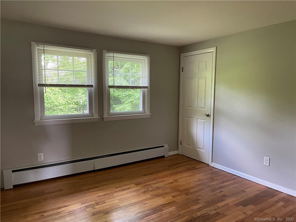 Empty bedroom with hardwood floors, two blinded windows, and a long baseboard heater.
