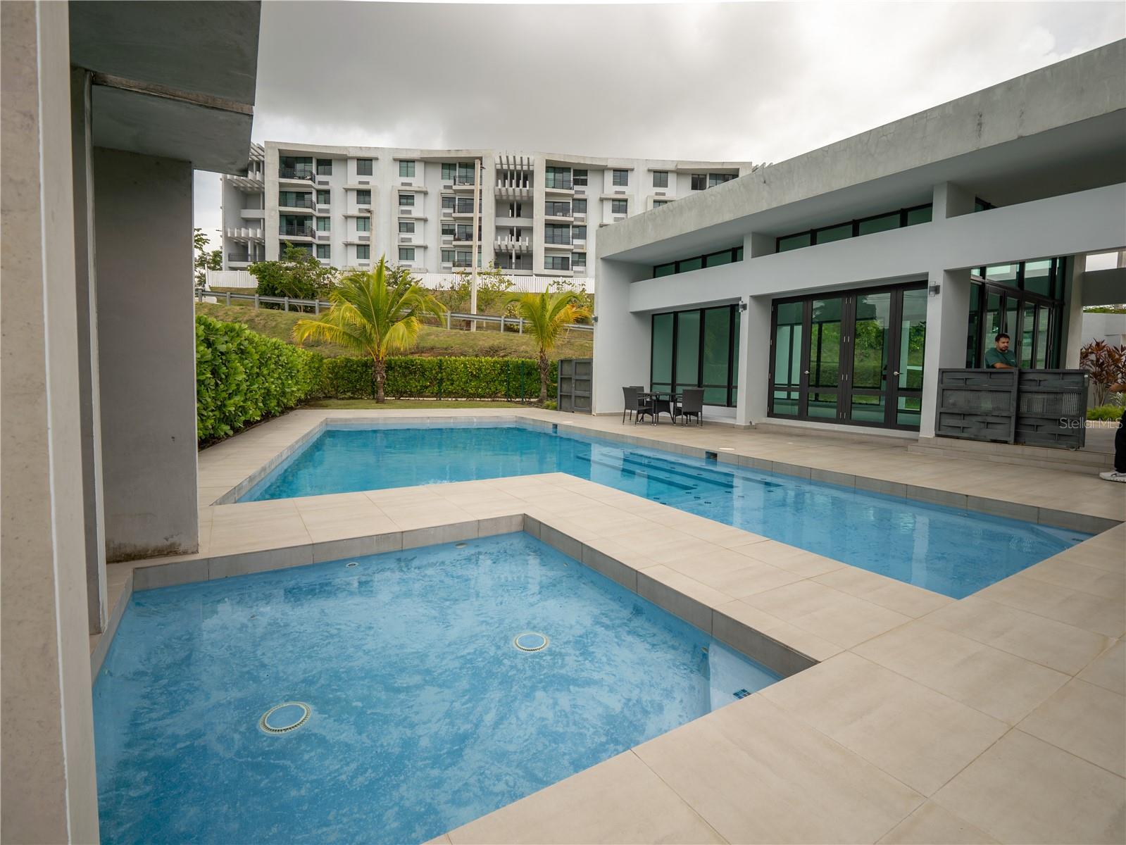 Two courtyard swimming pools beside a modern residential building with palm trees and patio seating.