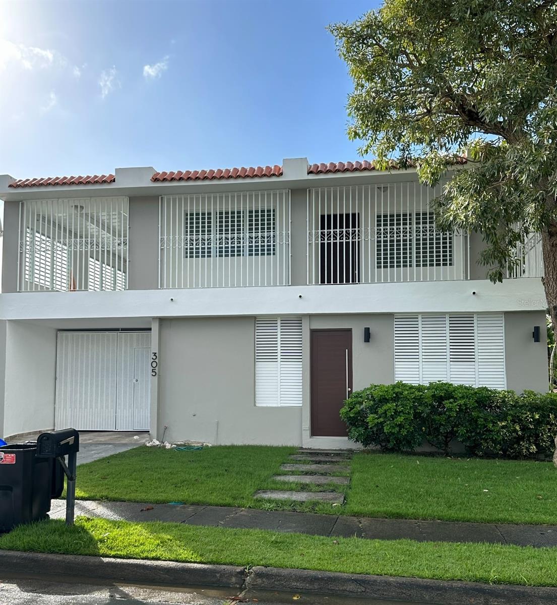 Two-story gray home with a red tile roof, barred balcony, white shutters, and a front lawn.